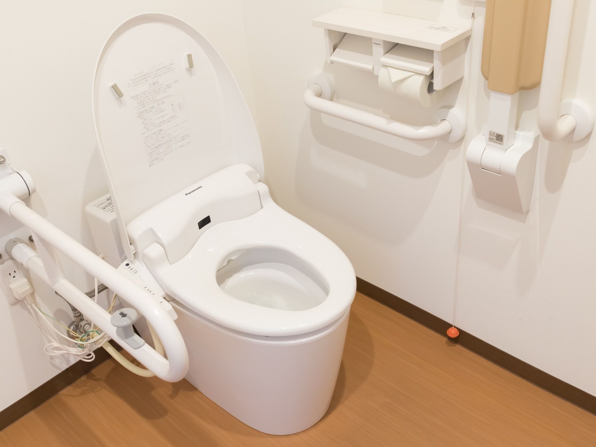 木目調の床に白い壁のトイレがある。便座の横には手すりが付いており、壁には長い紐のついたナースコールも備わっている。