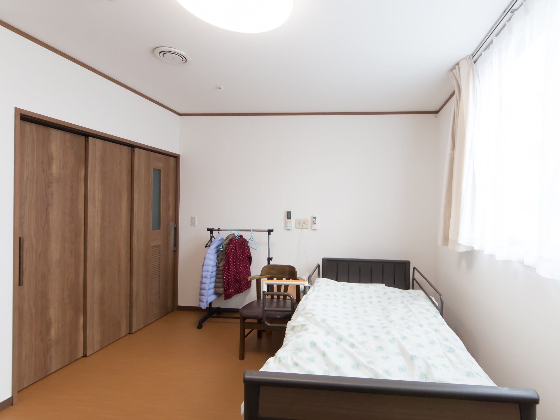 木目調の大きな引き戸が付いた居室がある。介護用ベッドが１台設置され、その横に椅子と衣装掛けが置かれている。