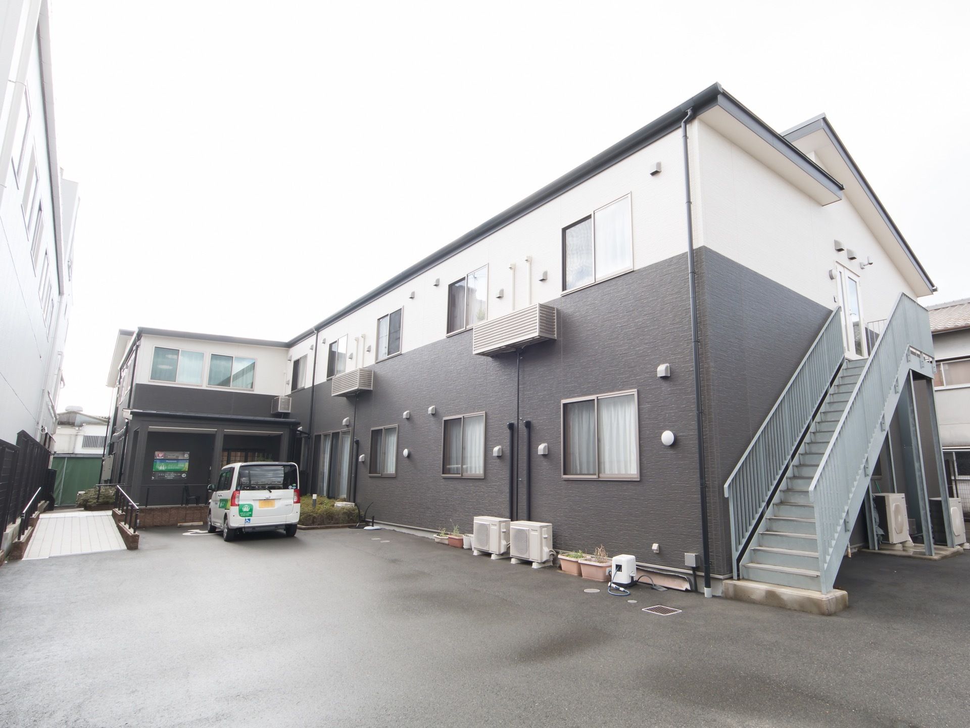 アスファルトで舗装された敷地に２階建ての住宅が建っている。玄関前は手すりの付いたスロープになっている。
