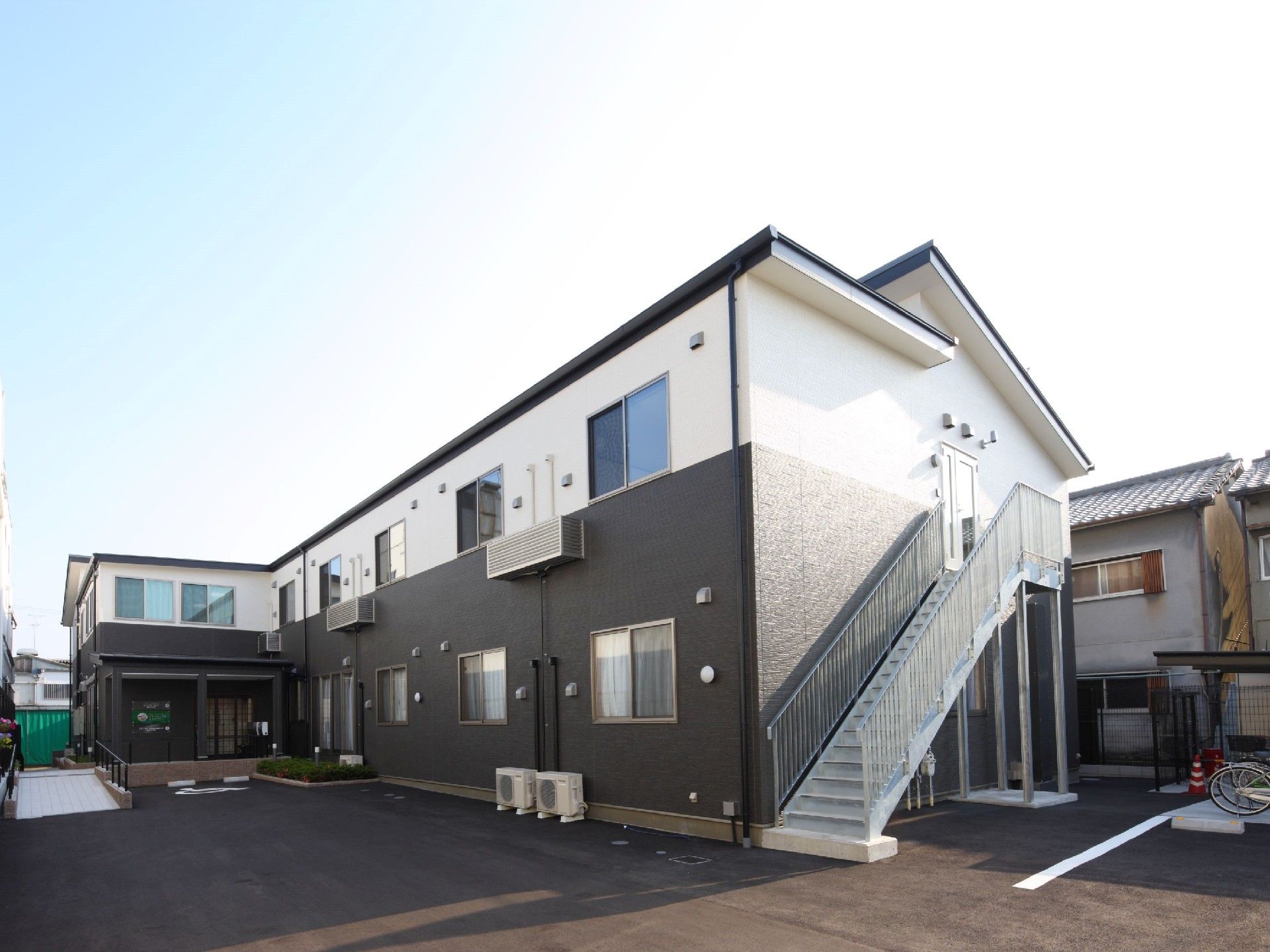 白と黒の2階建ての建物。玄関部分には屋根が付き、広い敷地内には駐車スペースもある。
