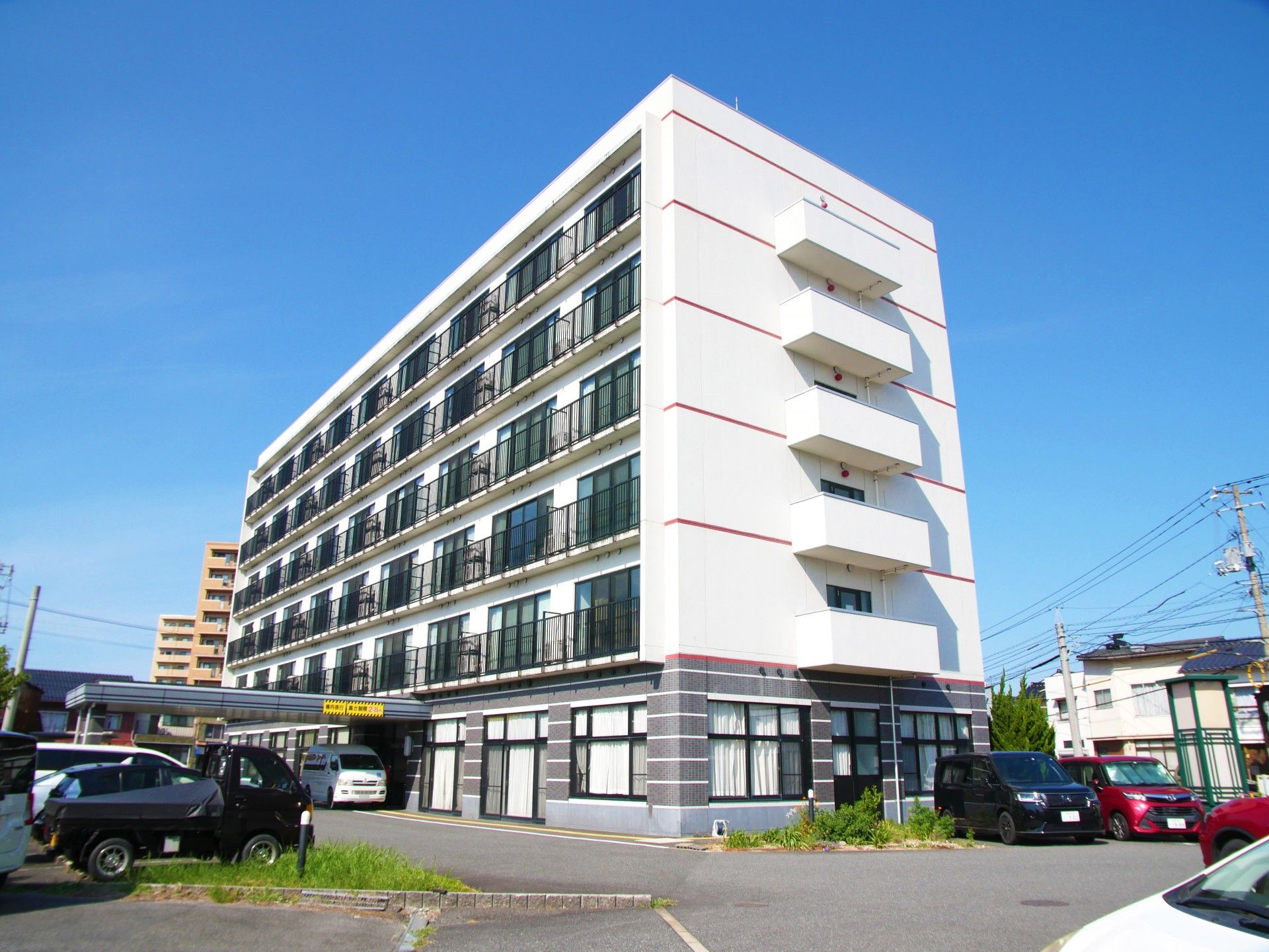 グレー系の大きな建物は各フロアにベランダがあり、玄関には車寄せが付き、周辺には住宅地がある。