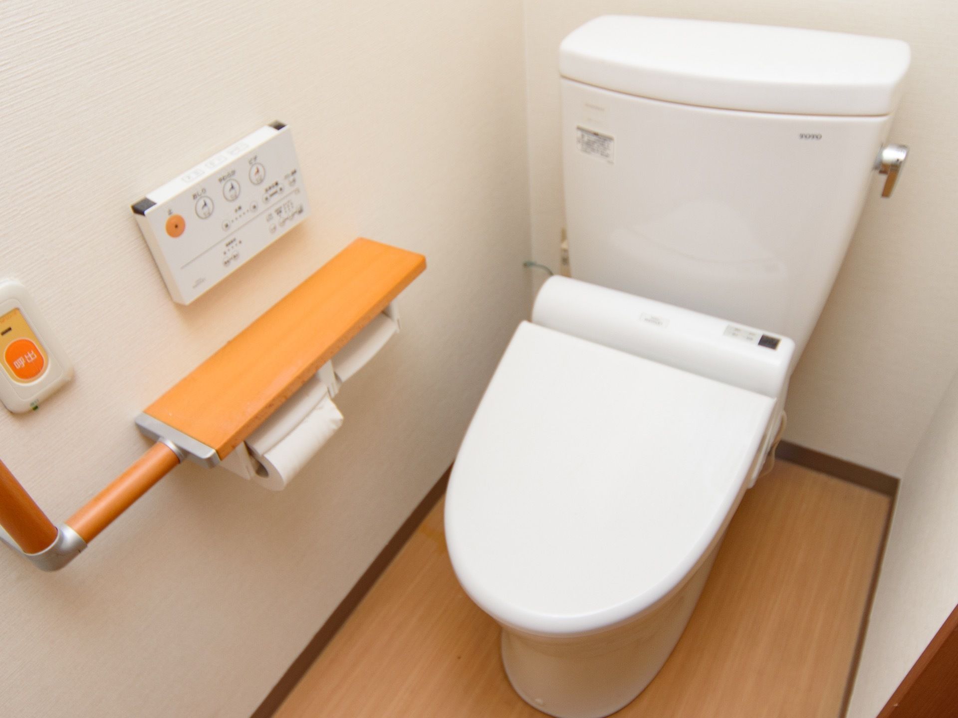 居室の完備されたバリアフリー設計のトイレである。温水洗浄便座や呼び出しボタン、手すりが壁に設置されている。