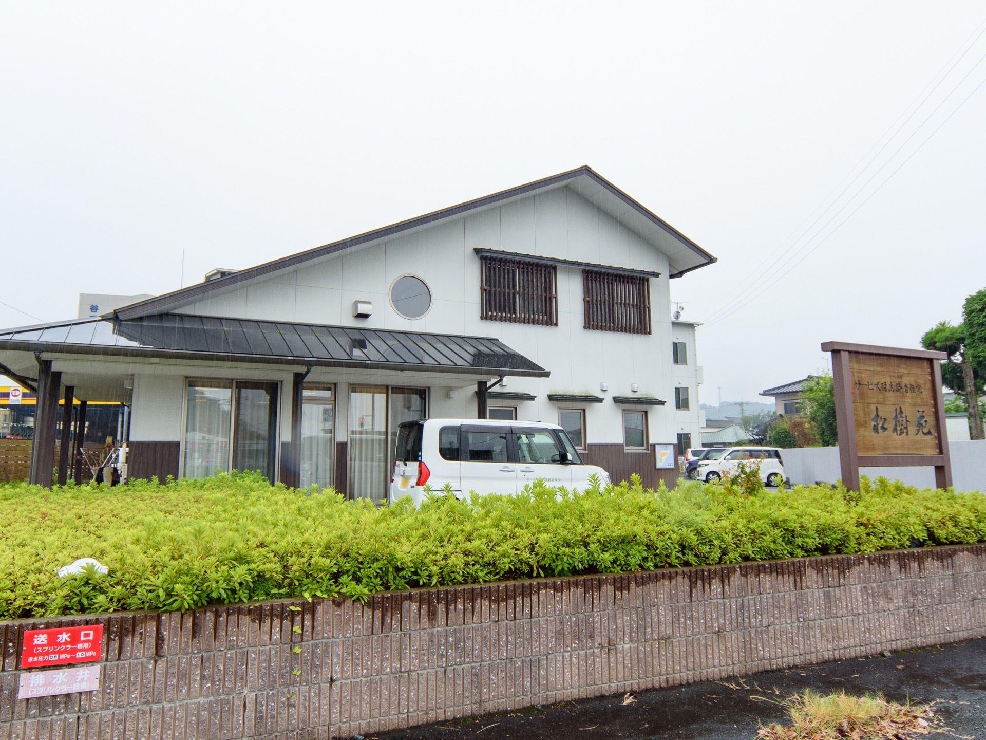 和風の外観の２階建て施設である。敷地内には、駐車場が用意されている。植え込みが設けられており、施設名が記された看板が設置されている。