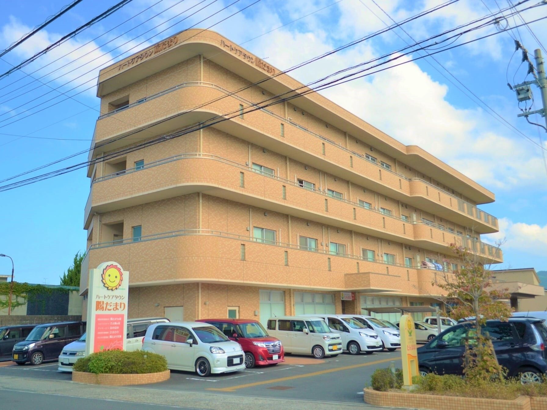 角フロアにベランダが付いた４階建ての建物の前には駐車場があり、たくさんの車が止まっている。