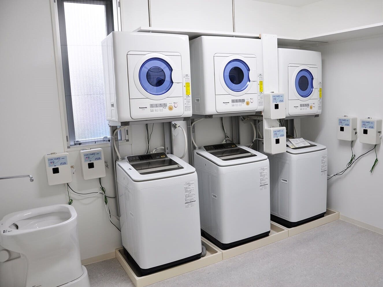 洗濯機置き場が３つ並んで設置され、それぞれ縦型の洗濯機が置かれ、その上には乾燥機も付いている。左側には手洗いスペースもある。