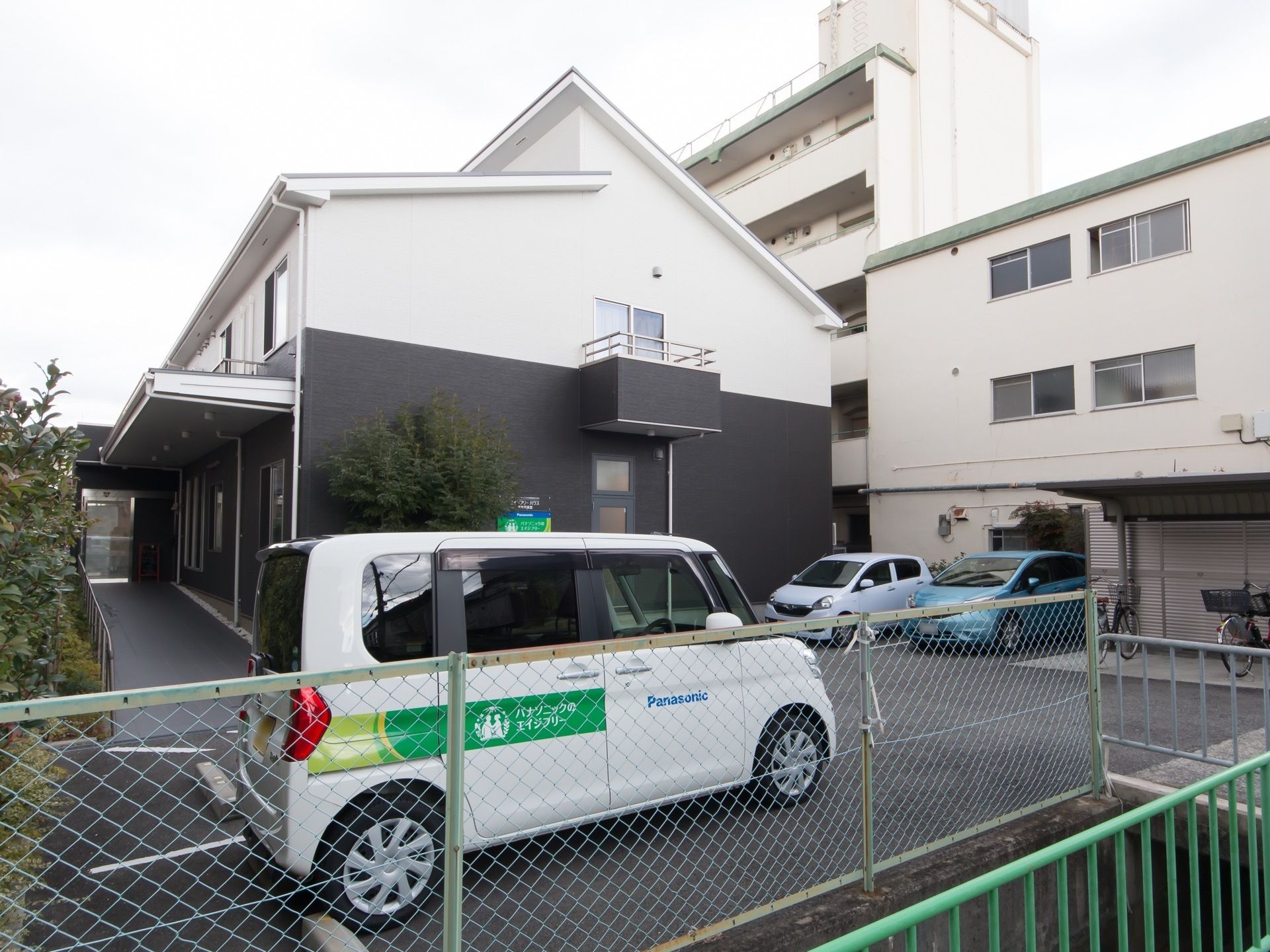 ２階建ての住宅が建っている。玄関アプローチは手すりの付いたスロープになっており、建物の前には車が数台停まっている。