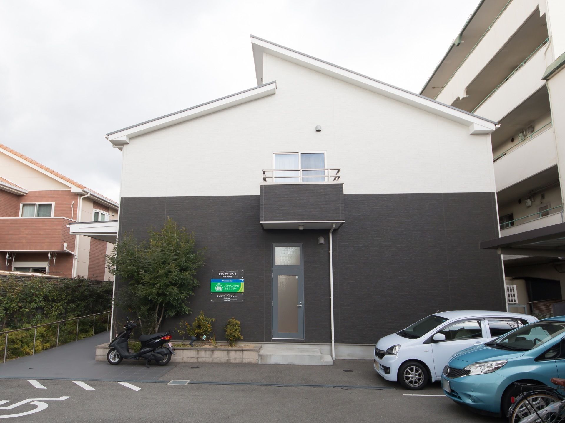 舗装された敷地の奥に、２階建ての住宅が建っている。手前は駐車スペースになっており、車が数台停められている。
