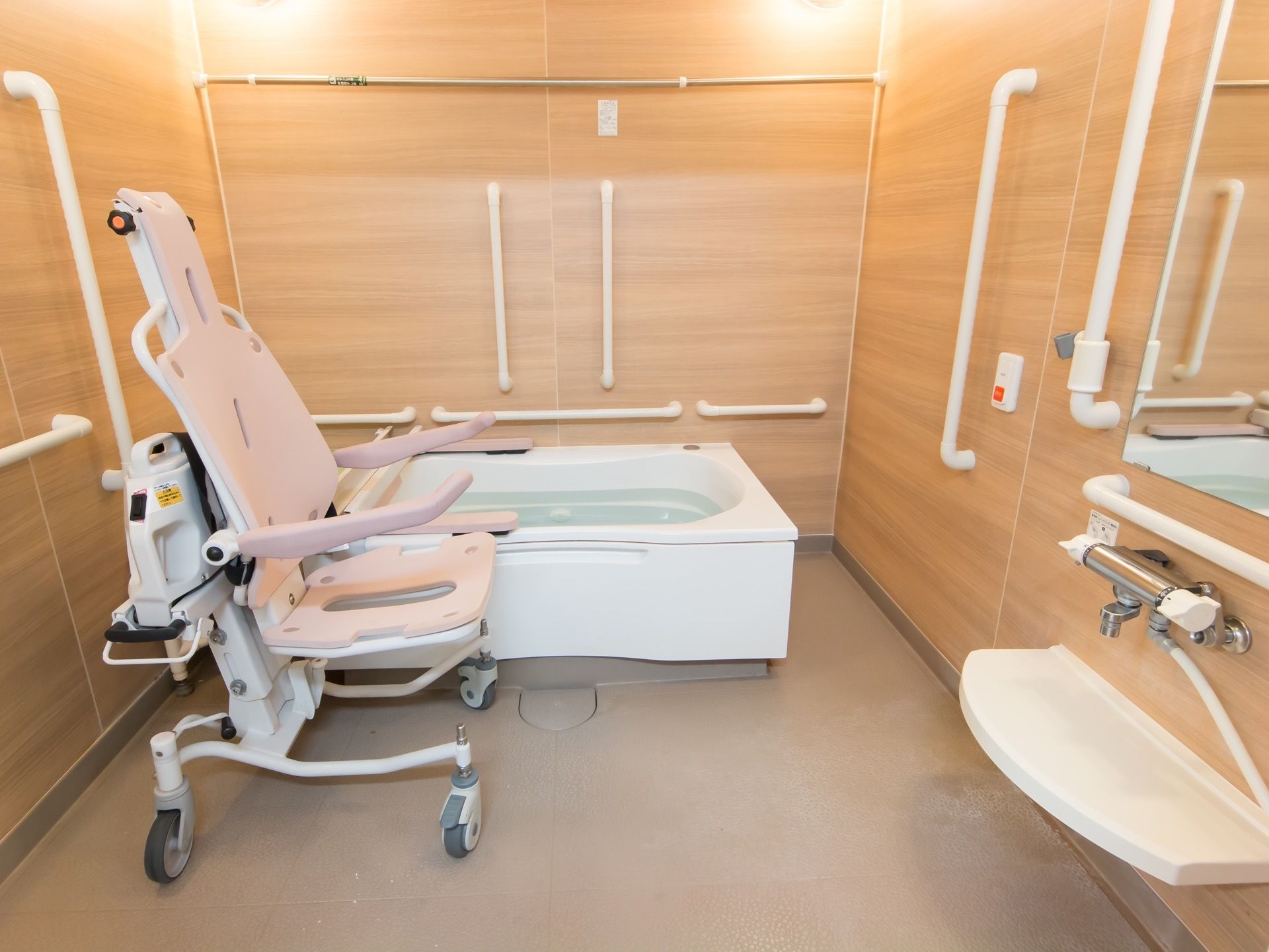 木目調の壁が印象的な広い浴室がある。介助用の車椅子が置かれ、浴槽の近くや洗い場の壁には手すりが備え付けられている。