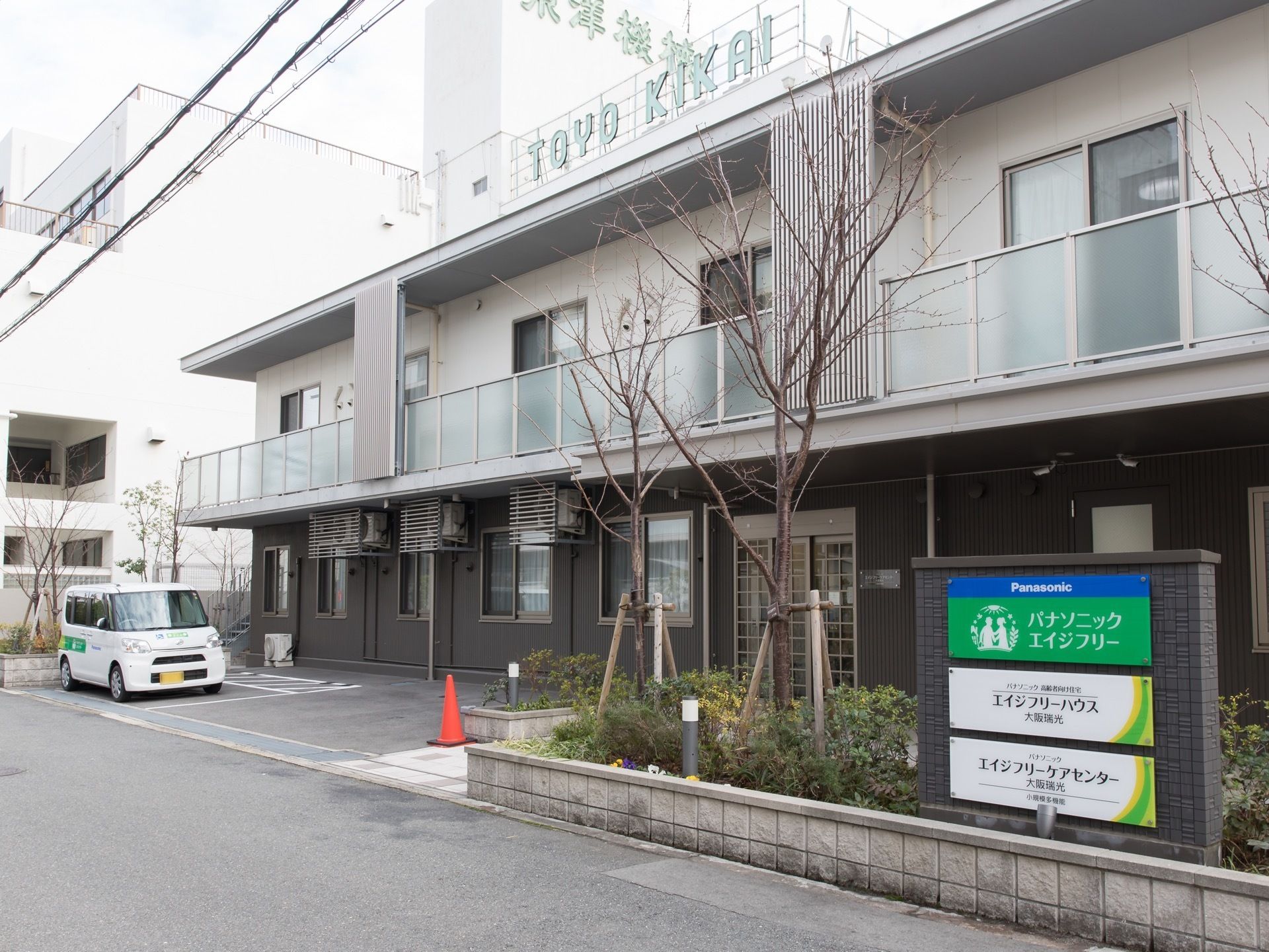 白とグレーのツートンカラにあしらわれた、２階建て施設である。庭木が植えられ、そばには看板が立っている。