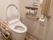 白色の洋式便座が備え付けられた、トイレである。可動式の手すりが、両サイドに設置されている。ナースコールもある。