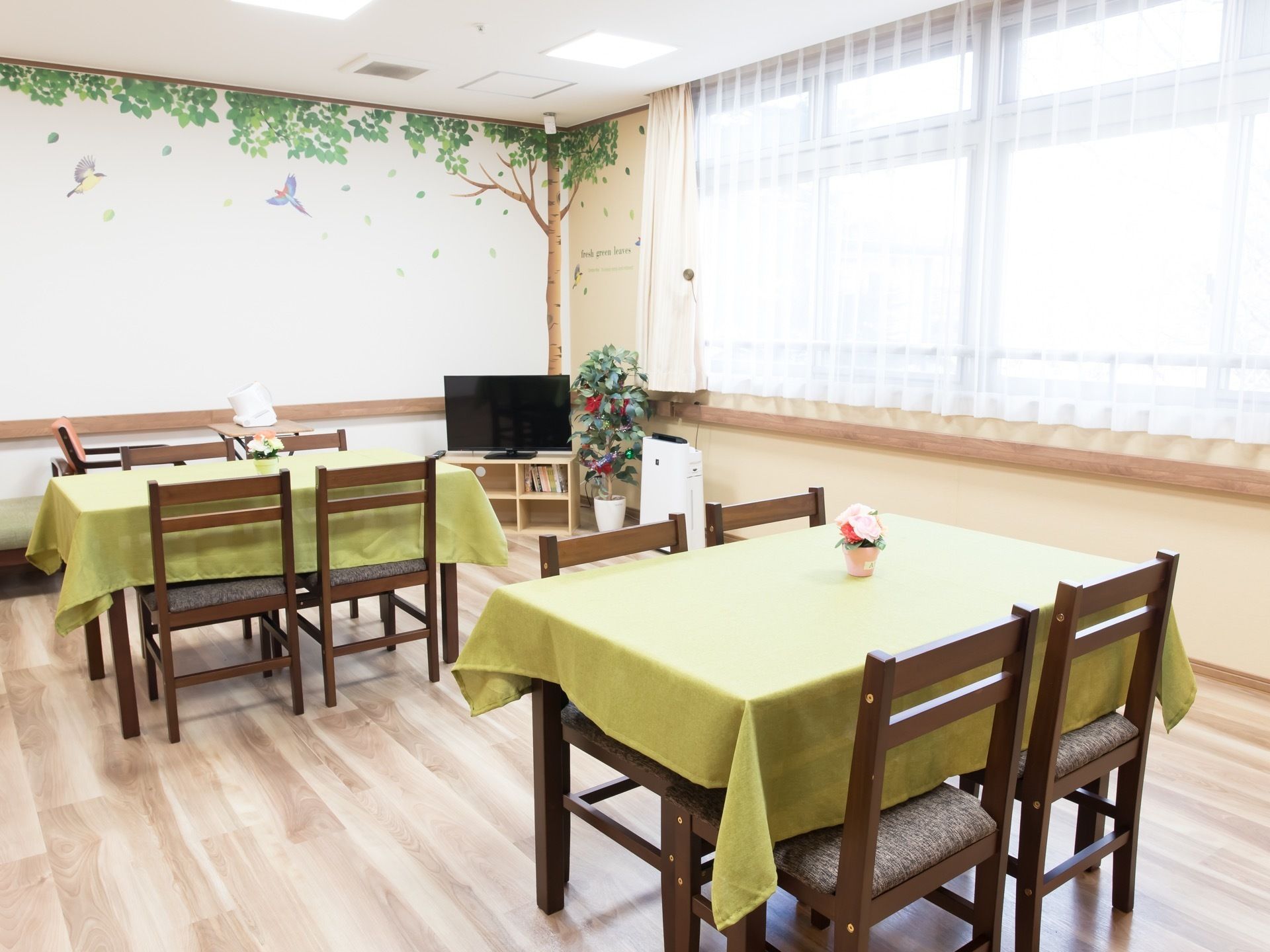 窓が大きく、光の差し込む食堂には、木調家具が使用され、あたたかな雰囲気となっている。壁には絵が描かれている。