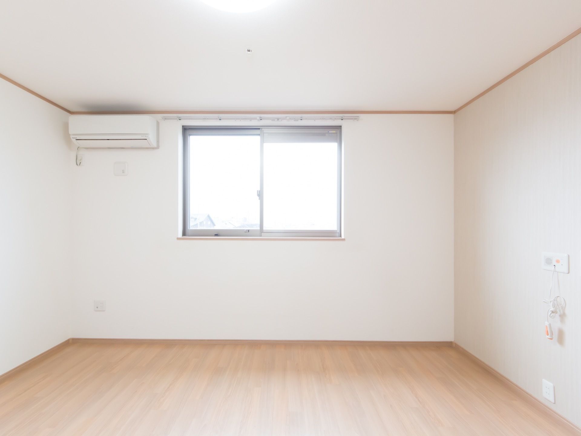 ベージュの床と白色の壁で清潔感ある広い居室。大きな窓があり明るい陽射しが入る。ナースコールとエアコンが設置されている。