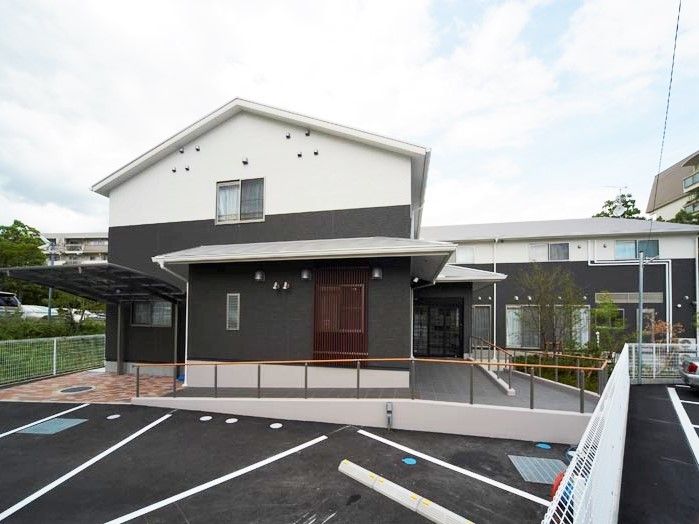 駐車場が付いた2階建ての建物には階段とスロープ付きの玄関がありり、周辺には緑もある。