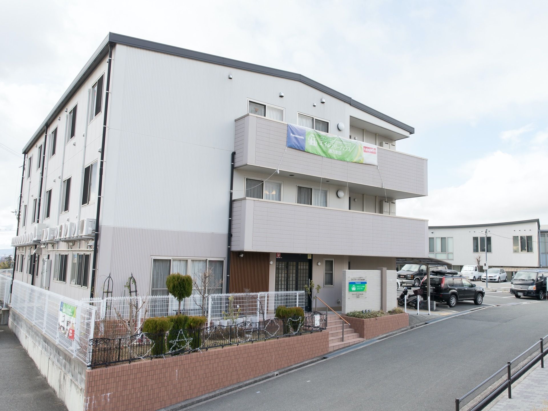 施設は３階建ての白い外壁の建物で、オシャレな集合住宅のように見える。柵にはさまざまな形の電飾が飾られている。