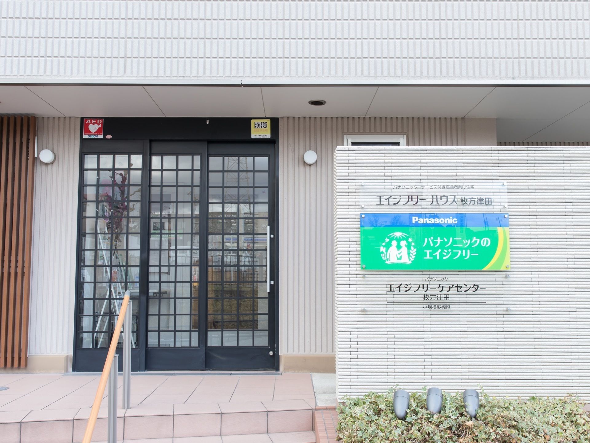 玄関ドアは、格子の入った３枚ガラスである。玄関前に階段があり、中央に手すりが付いている。玄関脇の白壁に、施設の名称が書かれている。