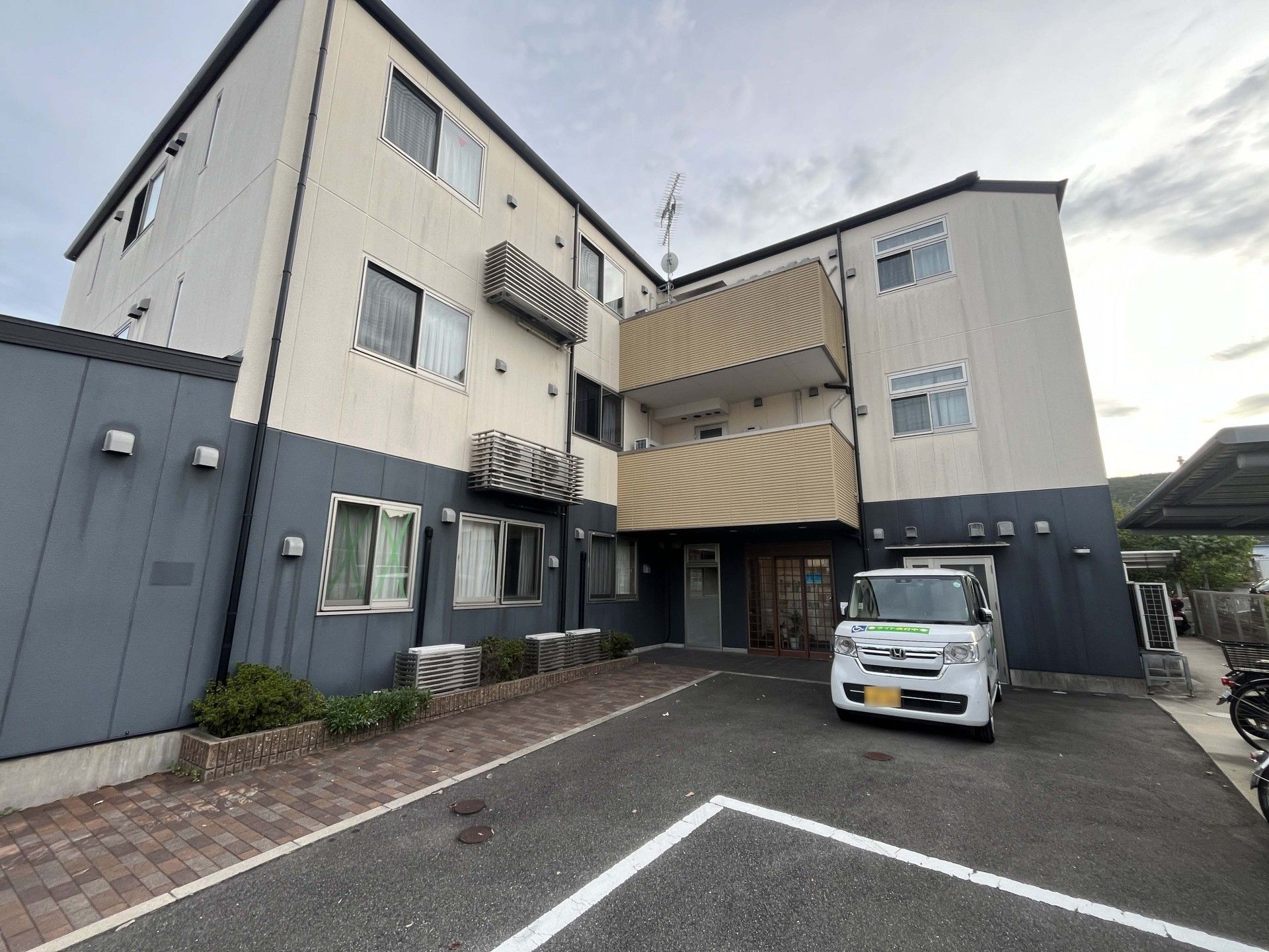 玄関には手すり付きのスロープが付いた3階建ての建物。駐車スペースもある。