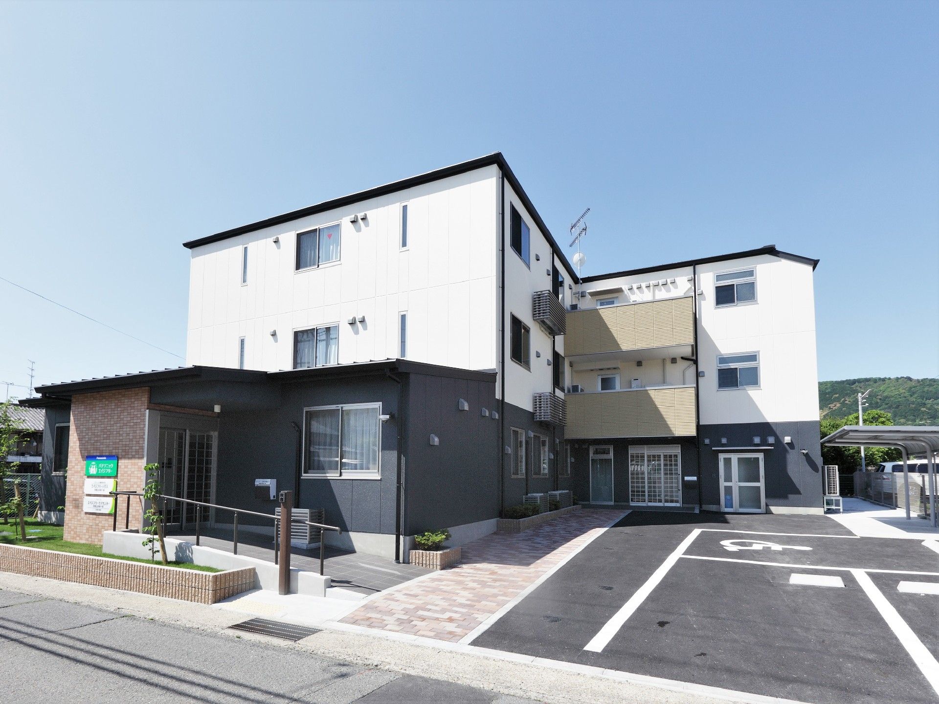 白とグレーの３階建ての建物。玄関先には手すり付きのスロープと屋根があり、駐車場もある。