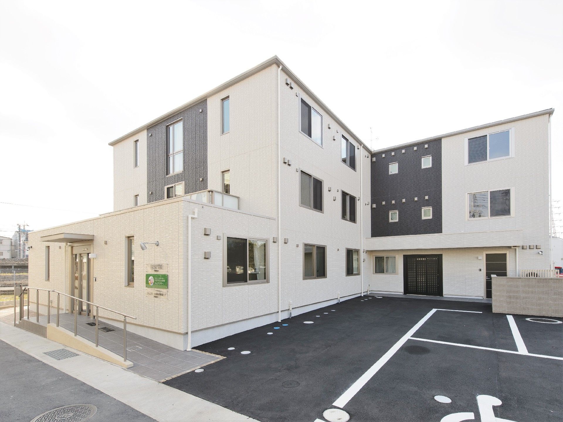 3階建ての建物は白をメインとしていて明るい雰囲気。玄関には屋根と手すり付きのスロープが付き、駐車場もある。