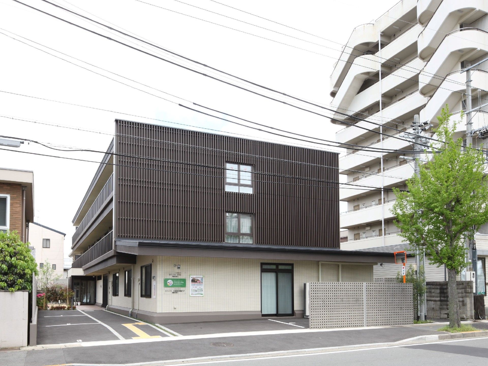 グレーと白の建物は3階建てで、ベランダや駐車場も設置され、周辺には住宅がある。