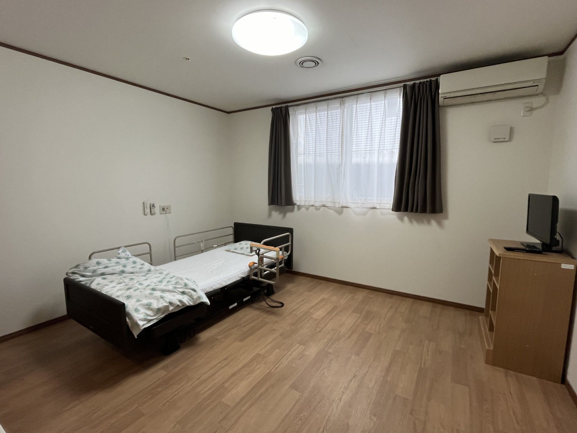 赤系のリーフ柄の寝具がセットされたベッドの横にはサイドテーブルがあり、トイレや洗面所がある居室。