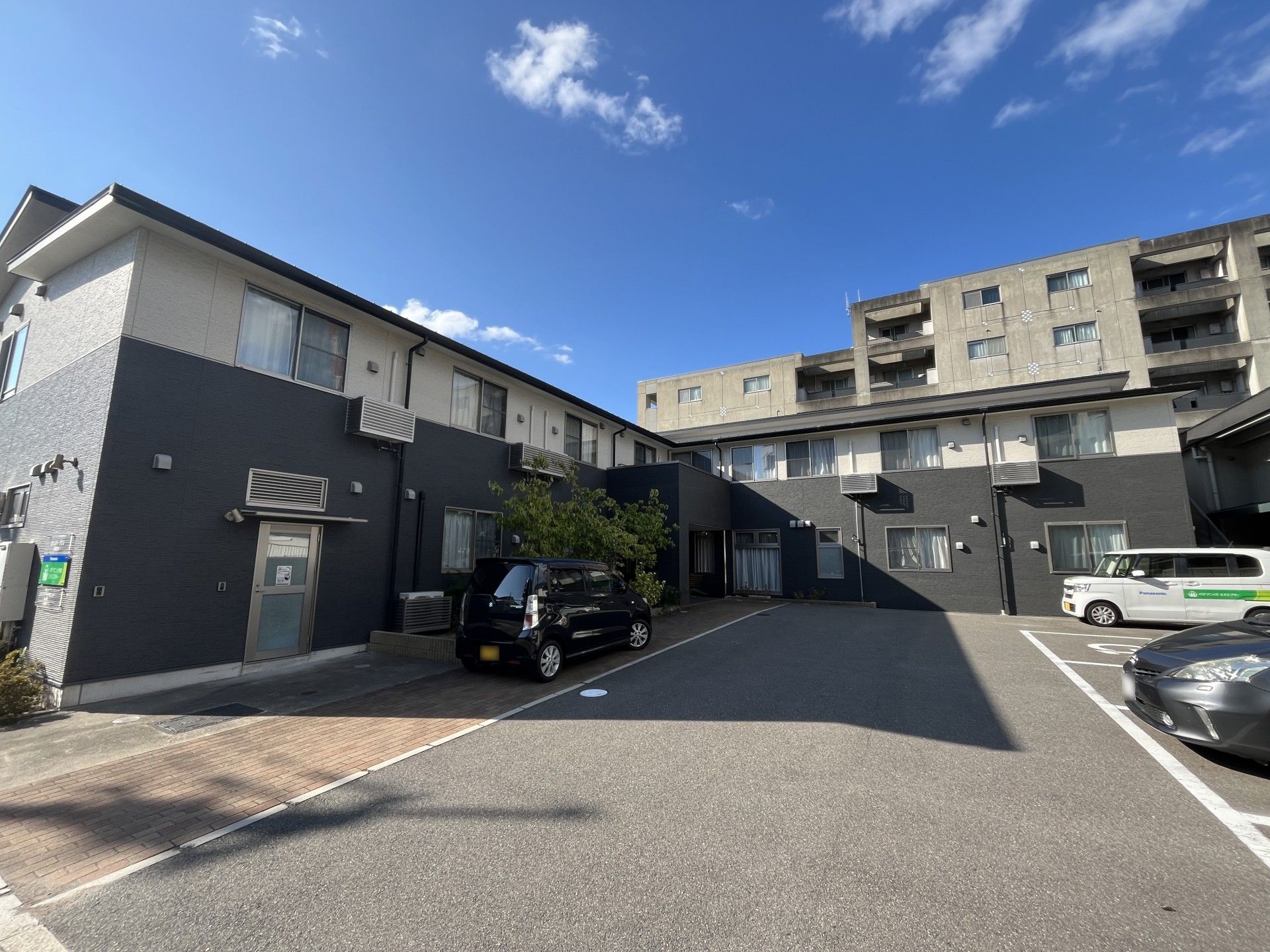白とダークグレーの2階建ての建物。玄関横には駐車場があり、外壁には住宅や併設事業所の名前が書かれたプレートが貼られている。