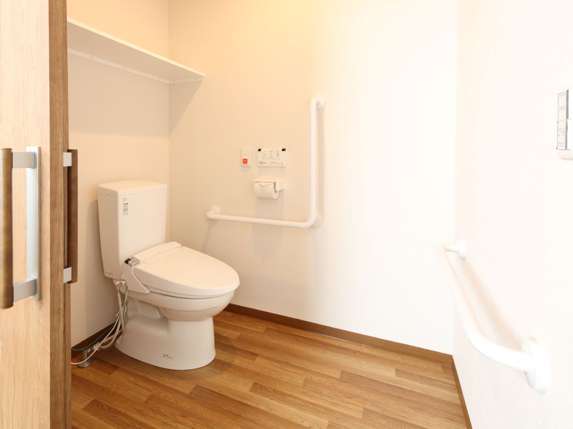 手すりや呼び出しボタンが付いたトイレはシンプルな造り。便座の上の方に棚もある。