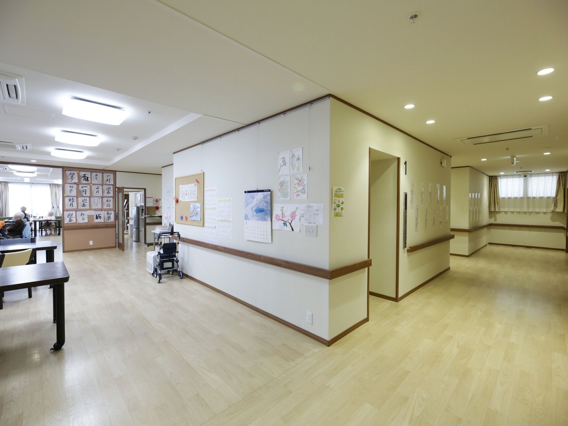 廊下との仕切りがない広間が食堂になっている。壁には板状の手すりがあり、入居者様の作品や掲示物が貼られている。