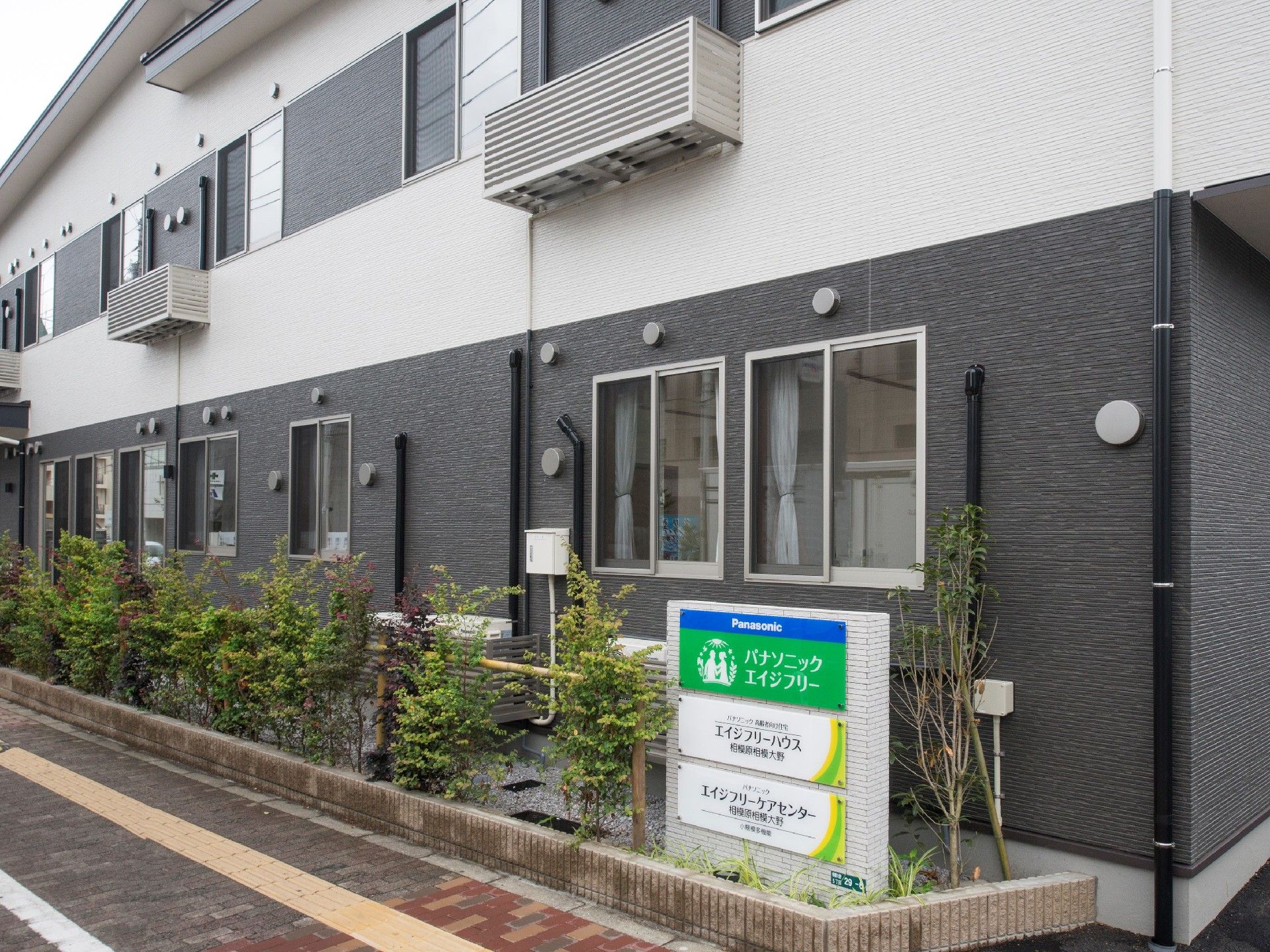 白とダークグレーの2階建ての建物。玄関先には看板があり、木も植えられている。