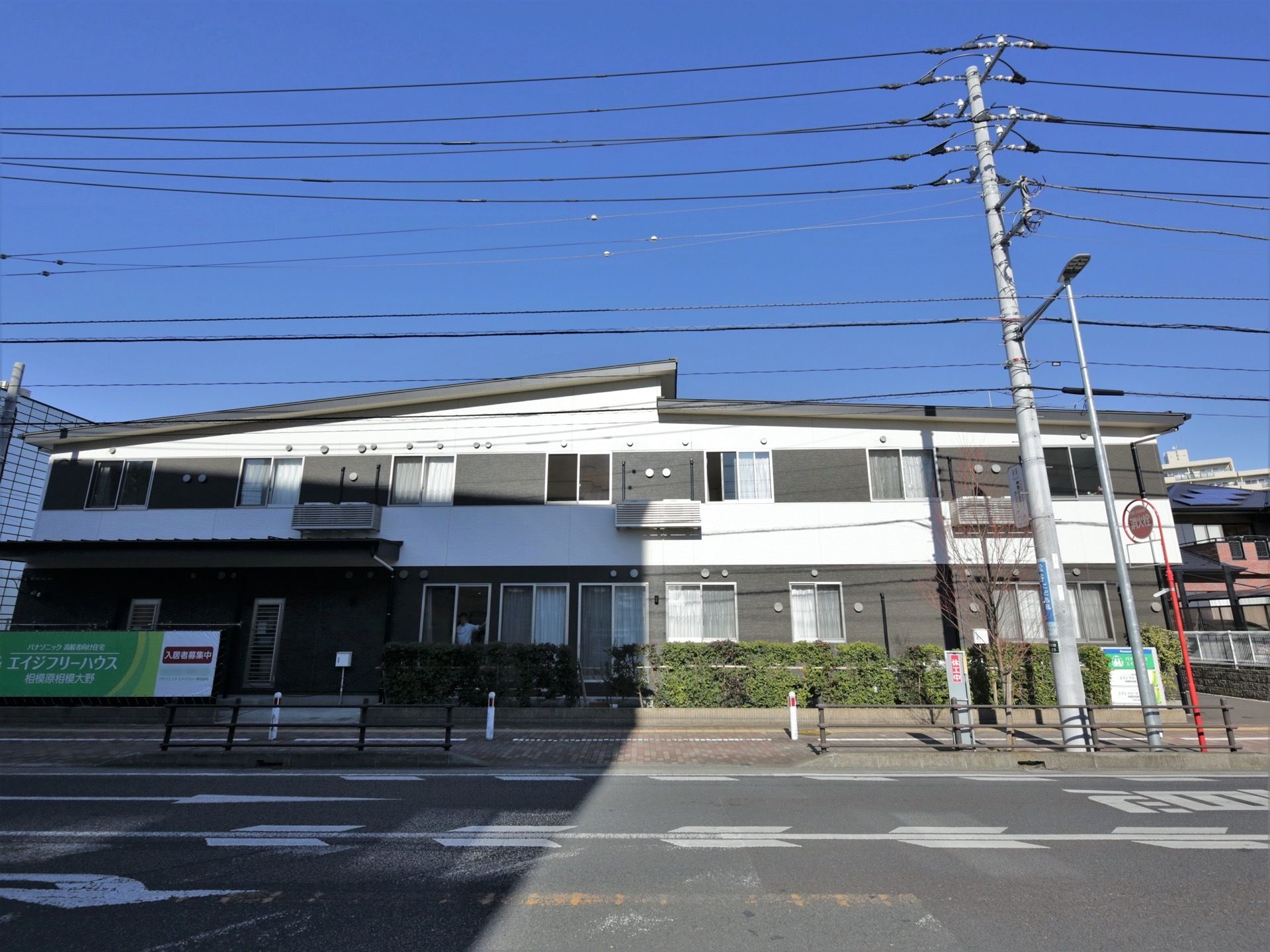 県道沿いに建つ２階建ての施設。白壁と黒壁のコントラストが目を引く。道路との間には植込みと施設の看板がある。