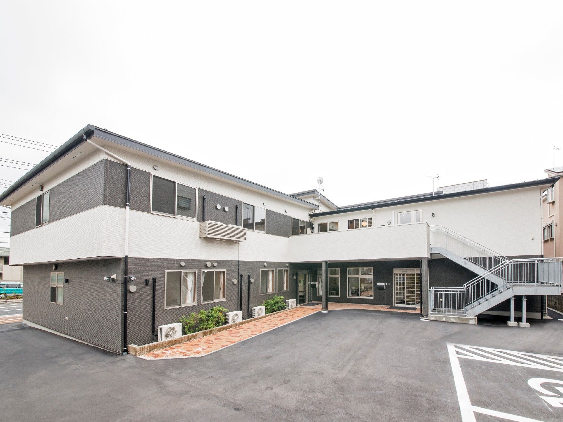 エル字型の２階建ての建物はモノトーン。建物周辺に緑が植えられ、駐車所が広い。