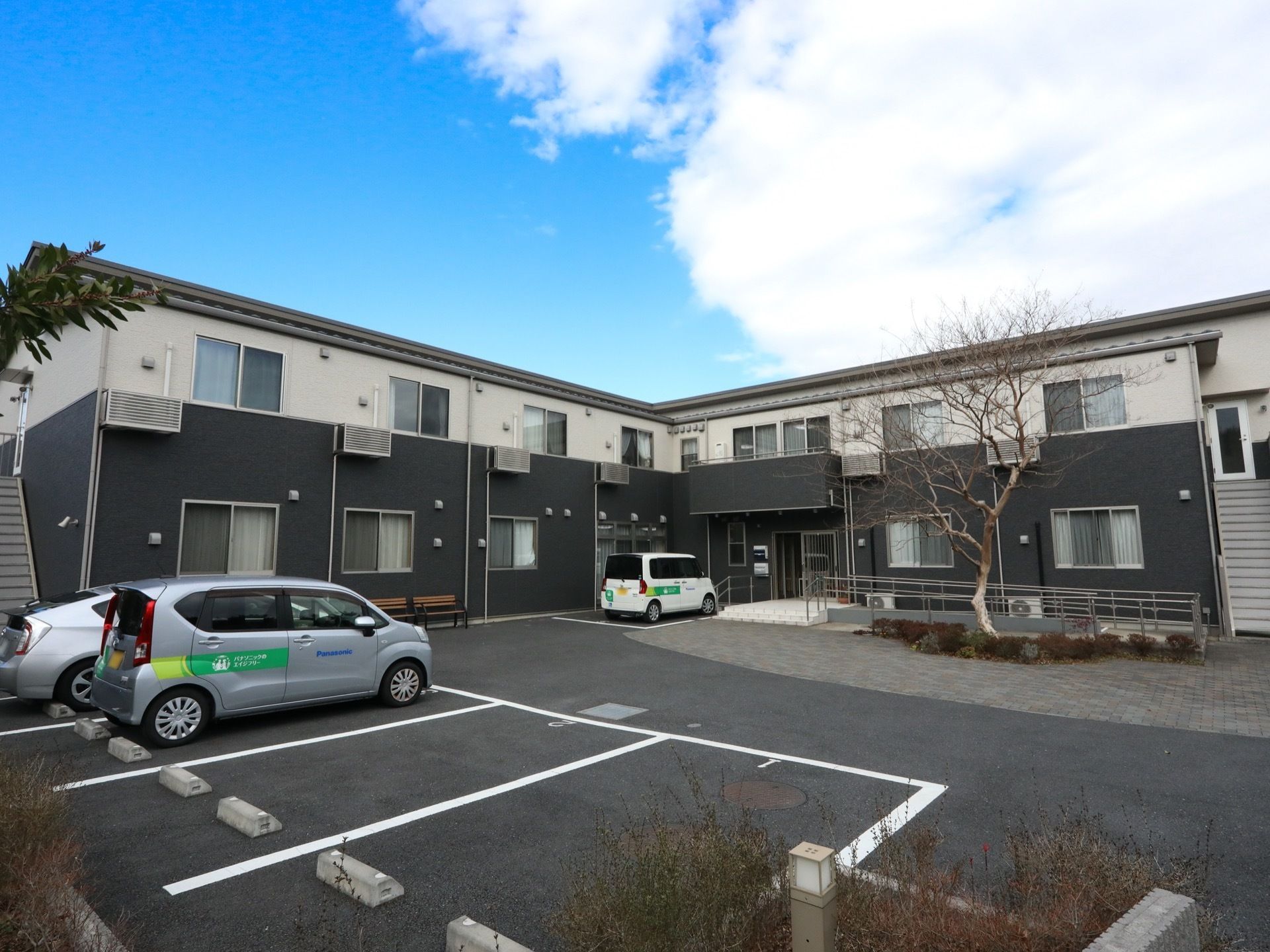 くの字型の構造をした２階建ての施設。玄関前に階段とスロープが設置されている。スロープの手前に背の高い木が生えている。