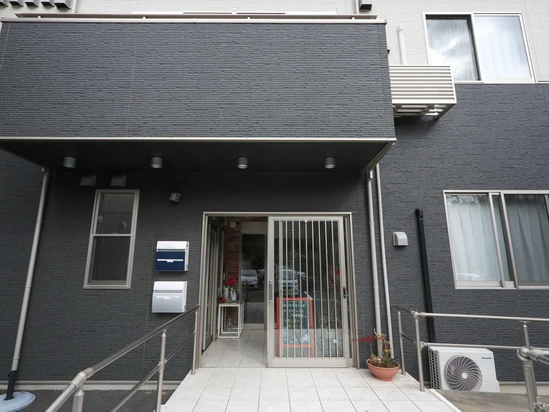 玄関の周囲は濃いグレーの外壁になっている。縦方向に筋が入ったガラスの引き戸である。左側の壁に郵便受けが２つ設置されている。