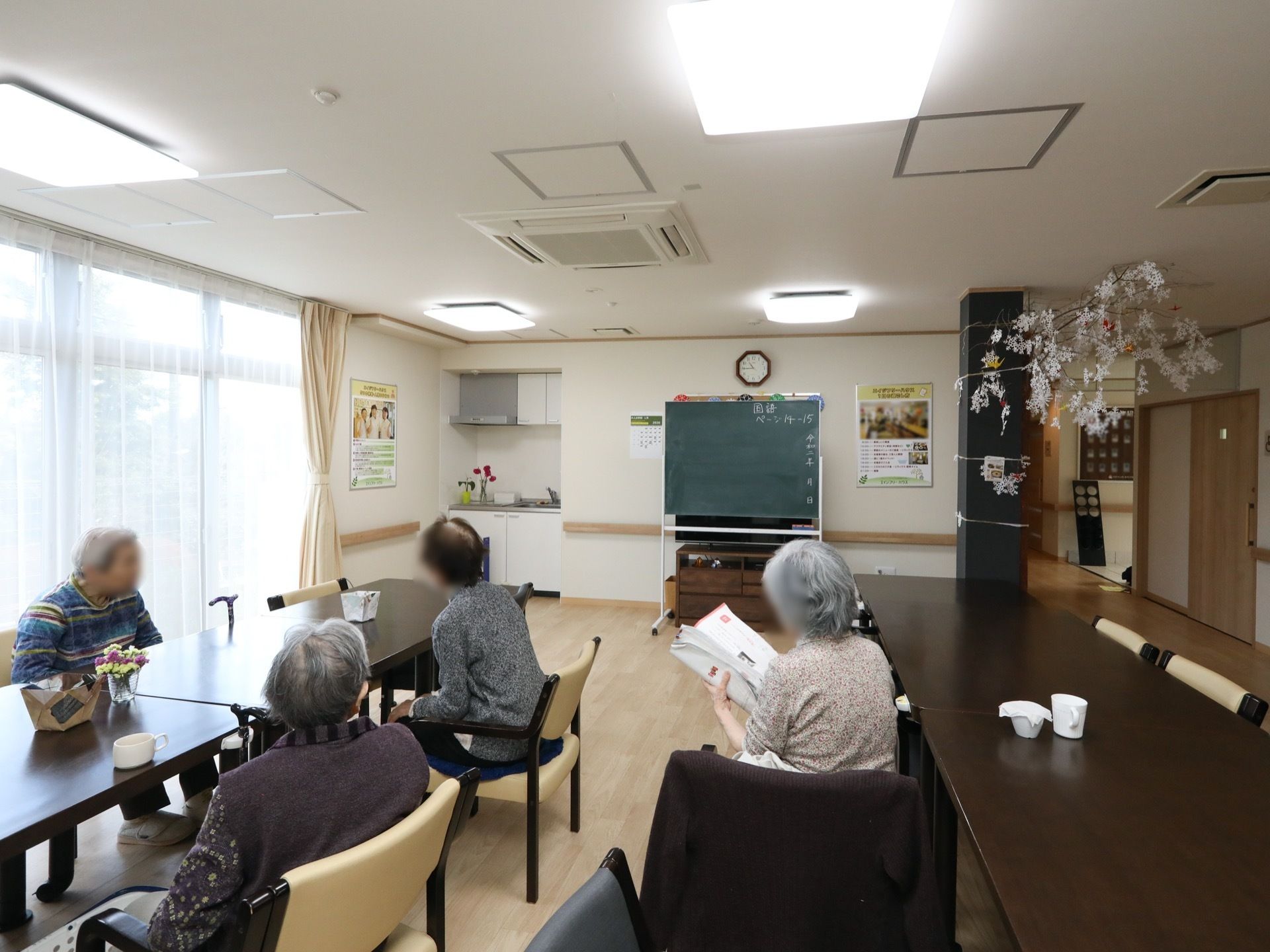 大きな掃き出し窓が並んでいる、明るい共有スペースがある。女性の入居者様が４名、集まって椅子に座っている。