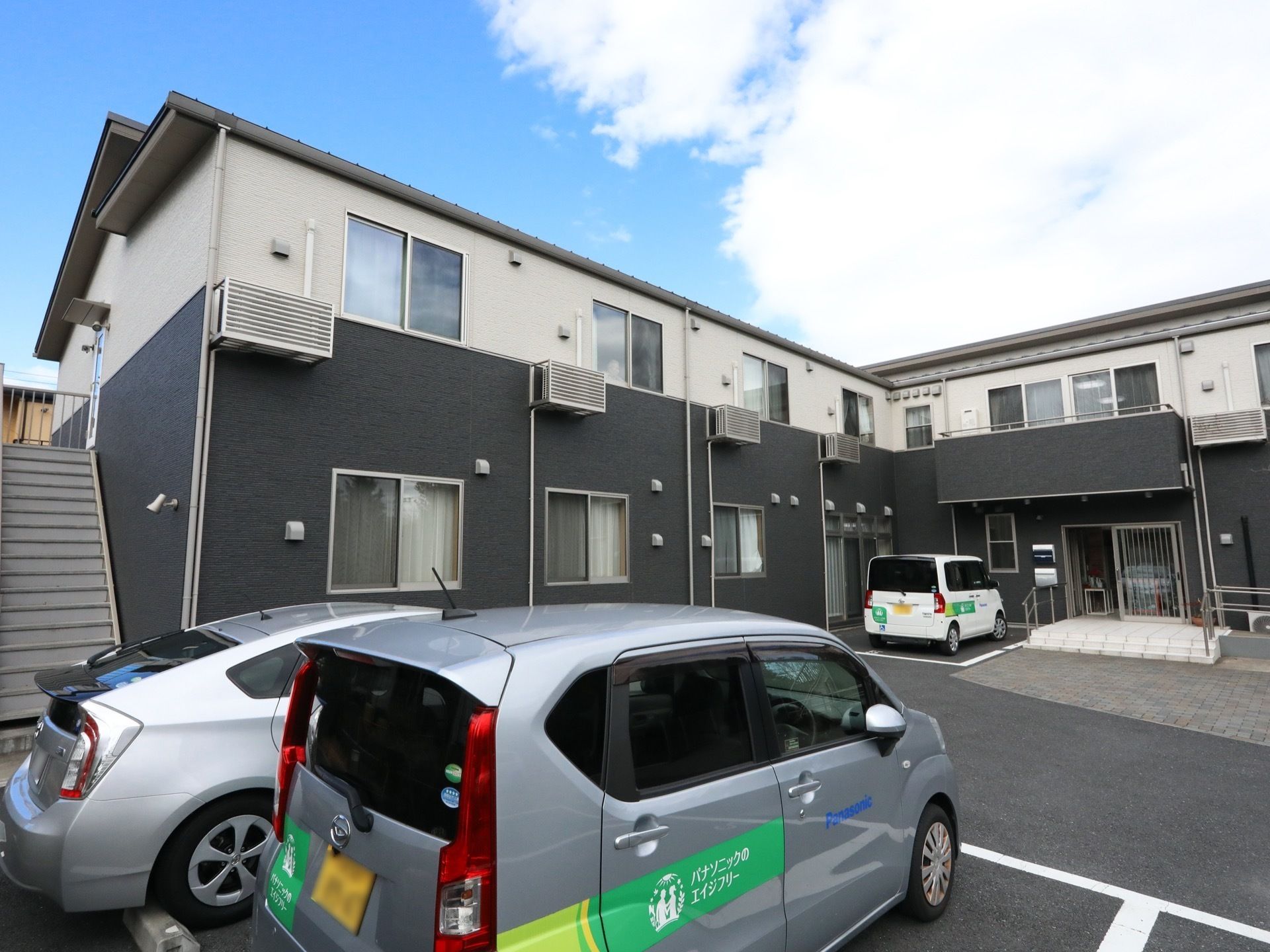 １階部分が濃いグレー、２階部分が白色のツートンカラーの外壁を持つ２階建ての施設である。施設の名称が表示された軽自動車が２台停まっている。