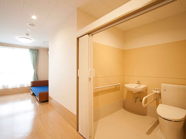 アコーディオンカーテンが付いた広いトイレが設置された居室。カーテンが付いた窓のそばには介護ベッドが置かれている。
