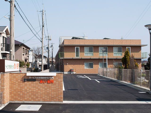 奥に茶色い建物と駐車場がある敷地入口にある施設名の書かれた塀