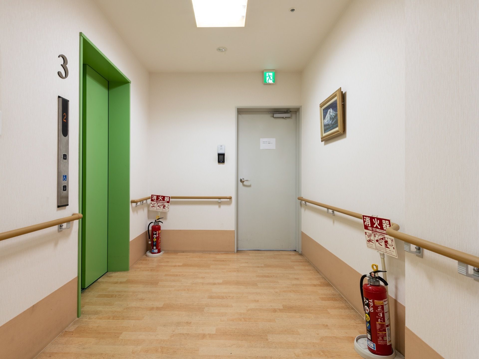 エレベータードアの正面の壁には絵画がかけられている。消火器が２つ置かれており、非常口の看板が設置されている。