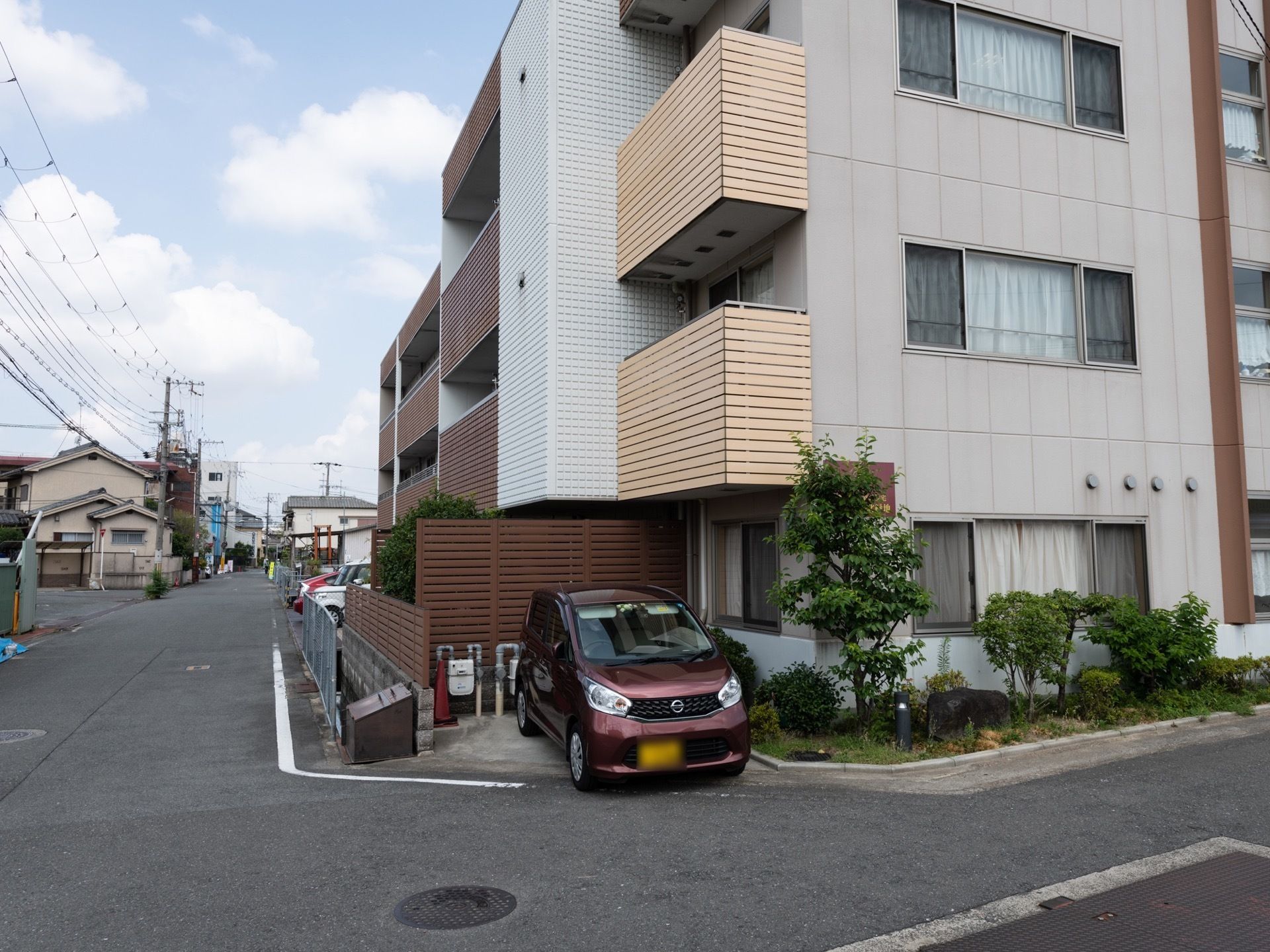落ち着いた色合いの施設外観となっている。1階には駐車スペースがあり、車が1台駐車されている。そばに植木が植えられている。