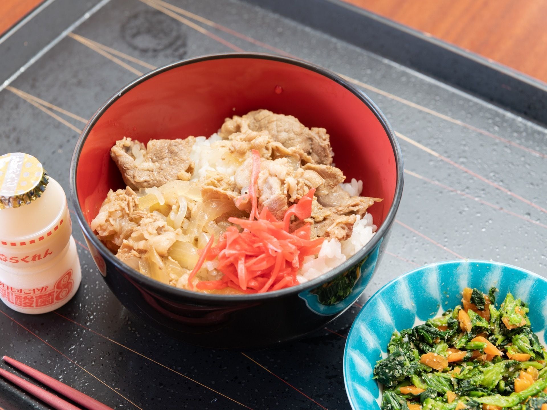 お盆の上に食事が用意されている。お盆には箸とほうれん草の胡麻和え、牛丼、乳酸菌飲料が置かれている。牛丼には紅しょうががかかっている。
