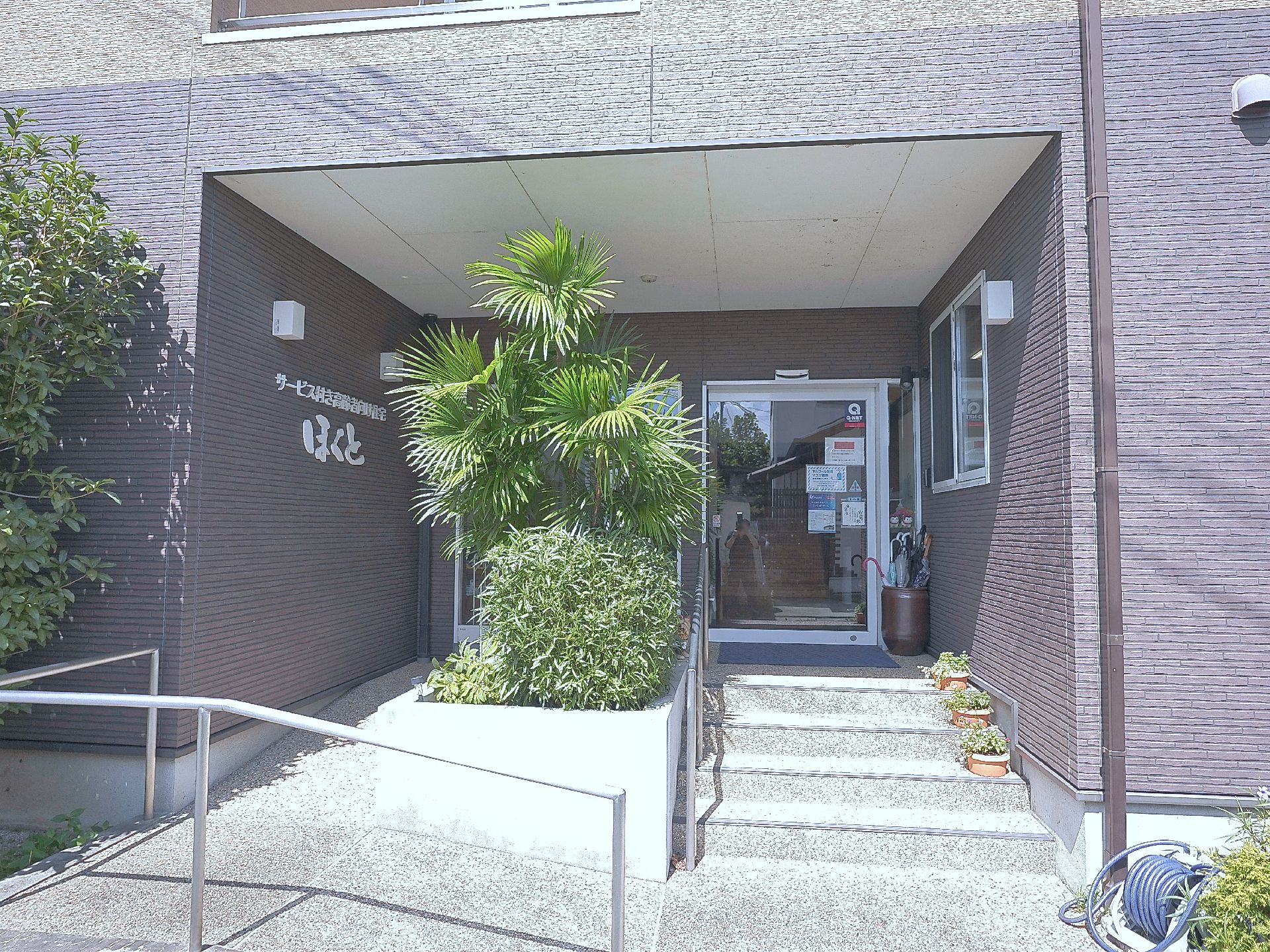 白いシャツを着た高齢男性がいすに座ろうとしているところを手伝っている若い女性スタッフ。ボーダー柄のエプロンを付け、髪を一つに結んでいる。