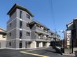 グレー系の建物は3階建てで、ベランダも付いている。広い敷地内には駐車場もあり、大きな看板も設置されている。