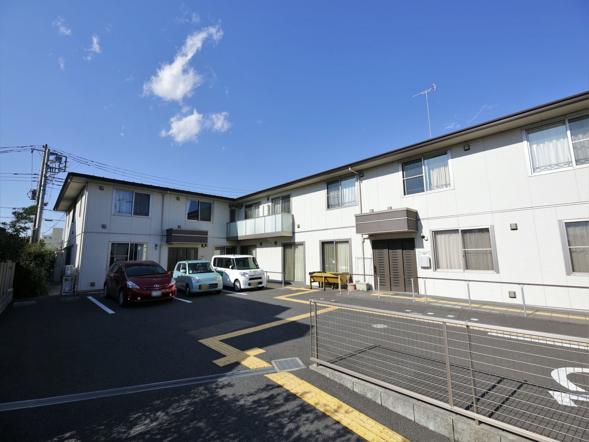 ２階建ての施設が右から正面に建っている。居室の窓が並んでいる。施設の前は駐車場になっており、車が３台停まっている。