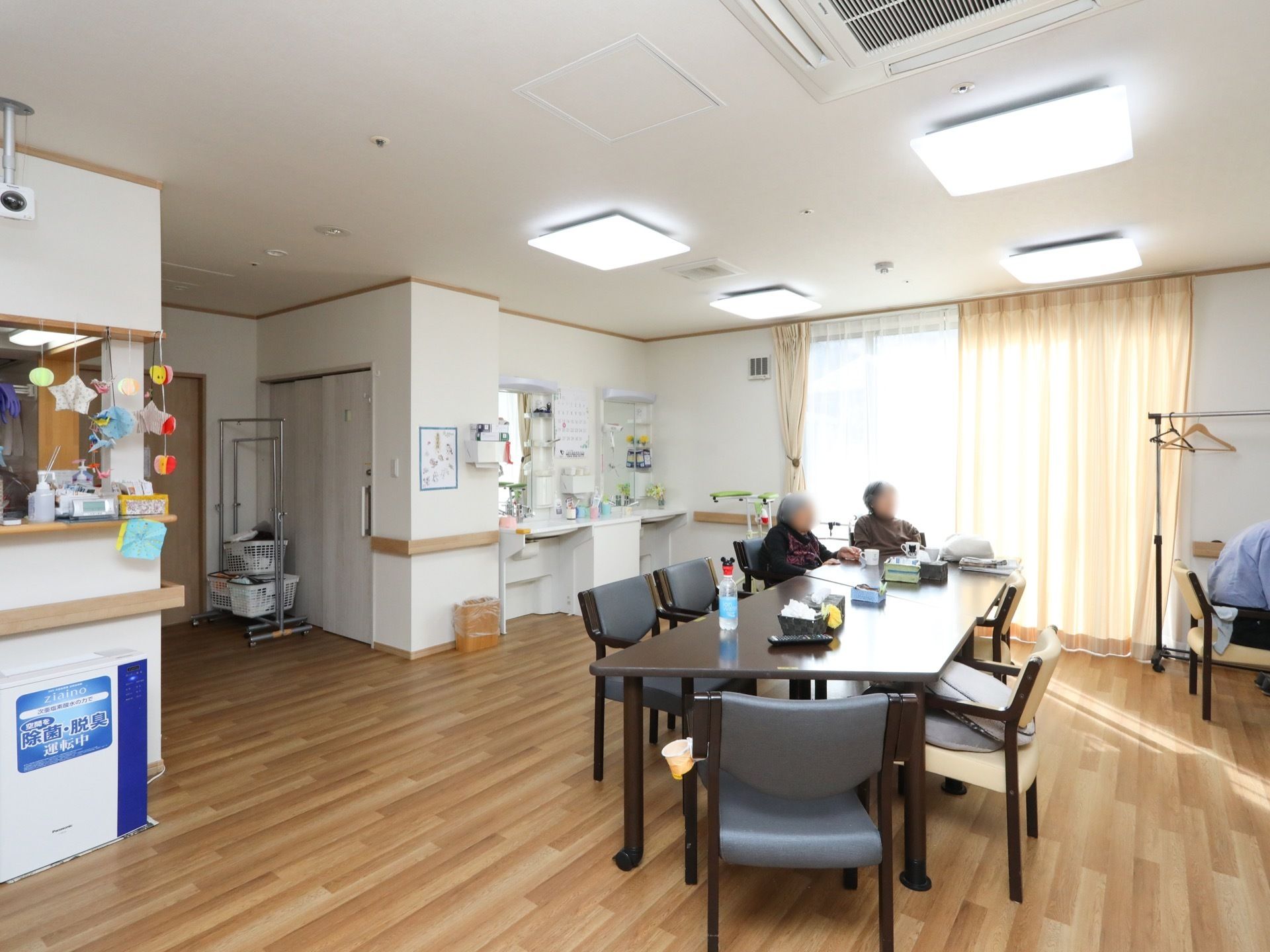 食堂に、台形の天板がついたテーブルが２つ組み合わせて置かれている。女性の入居者様が２名、並んで座っている。