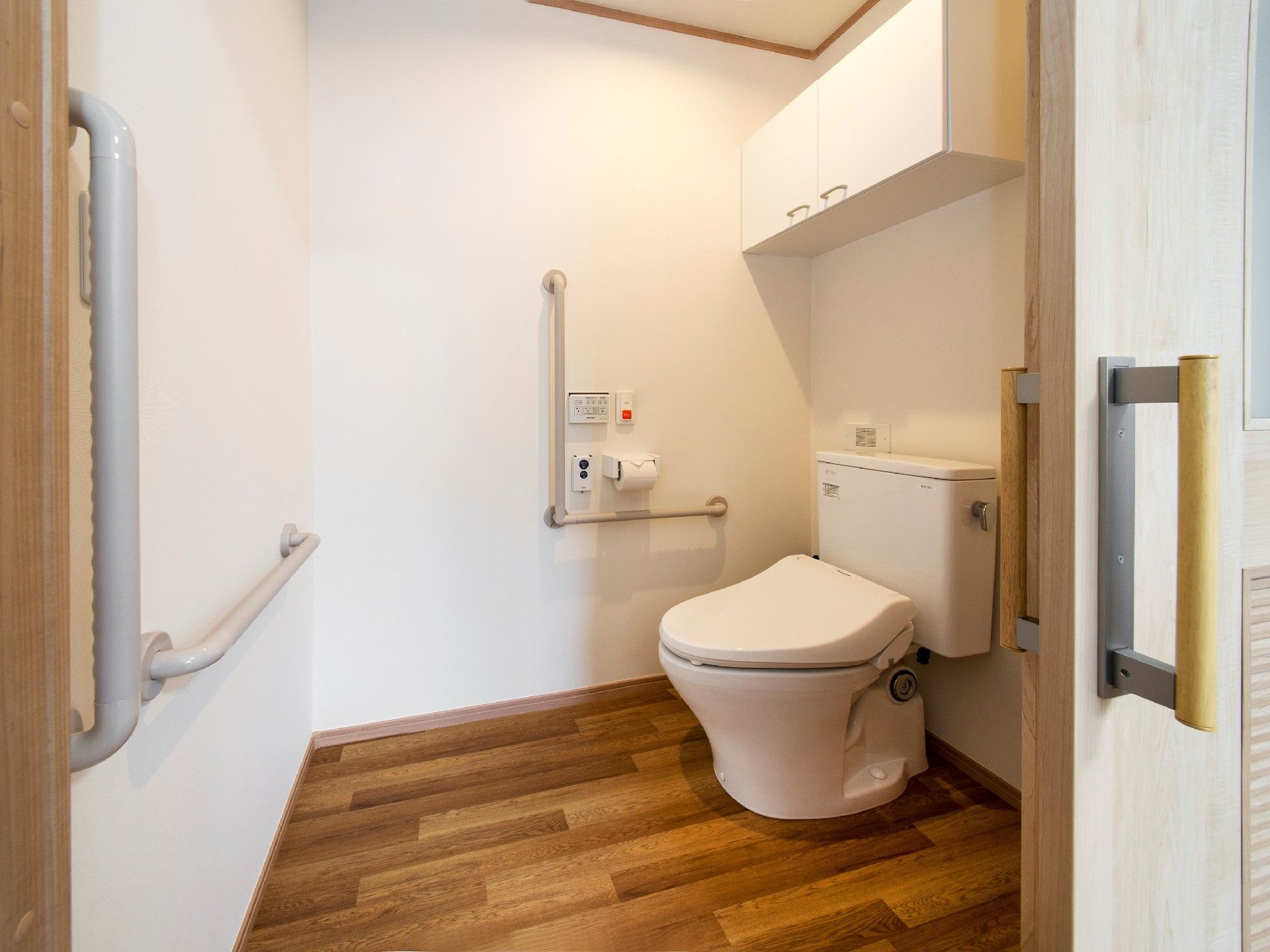 引き戸が付いたトイレは広く、壁にエル字型の手すりと呼び出しボタン、扉付きの棚が付いている。