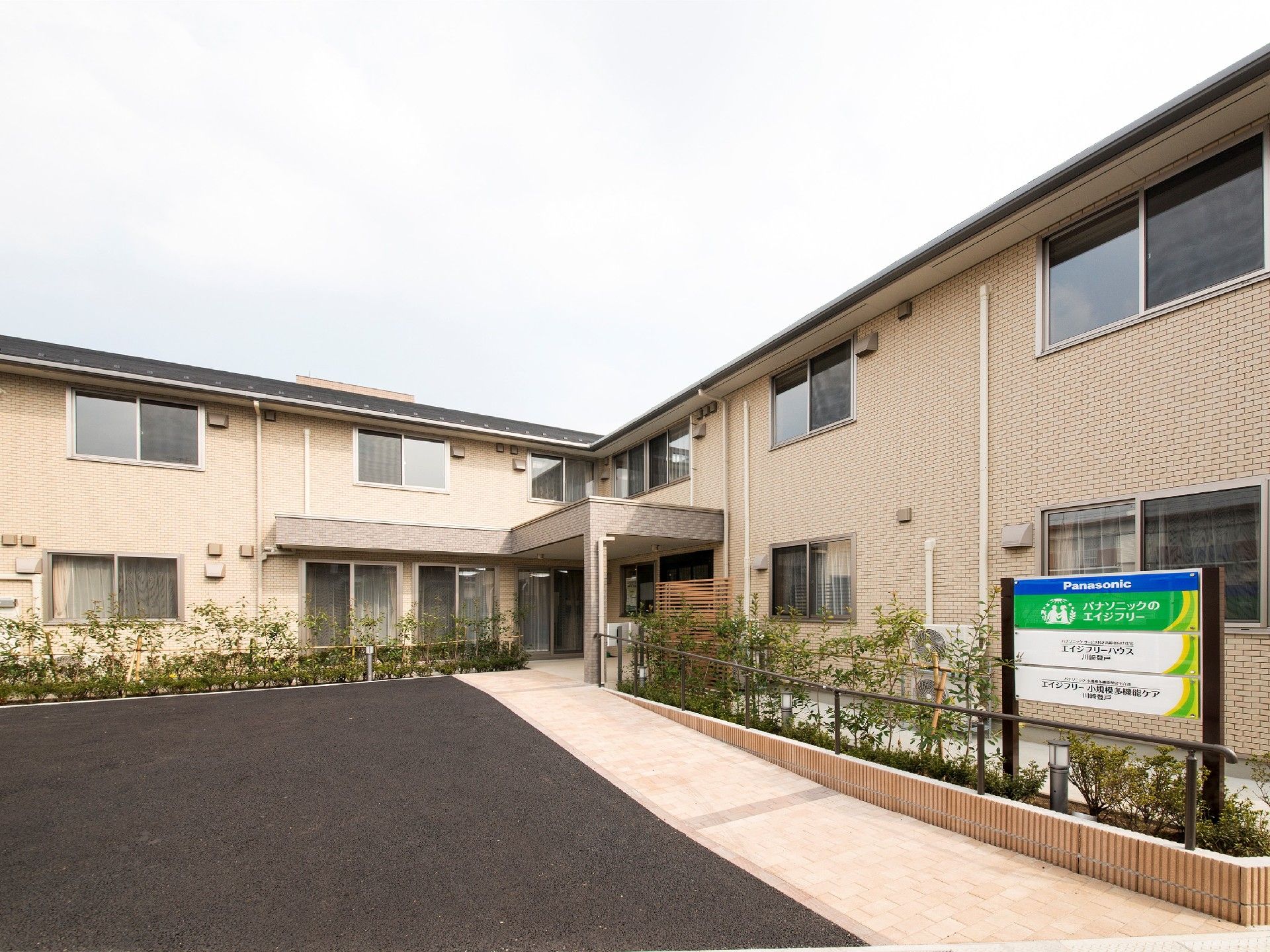 ベージュの2階建ての建物はエル字型で玄関には屋根が付き、建物周辺には木が植えられている。