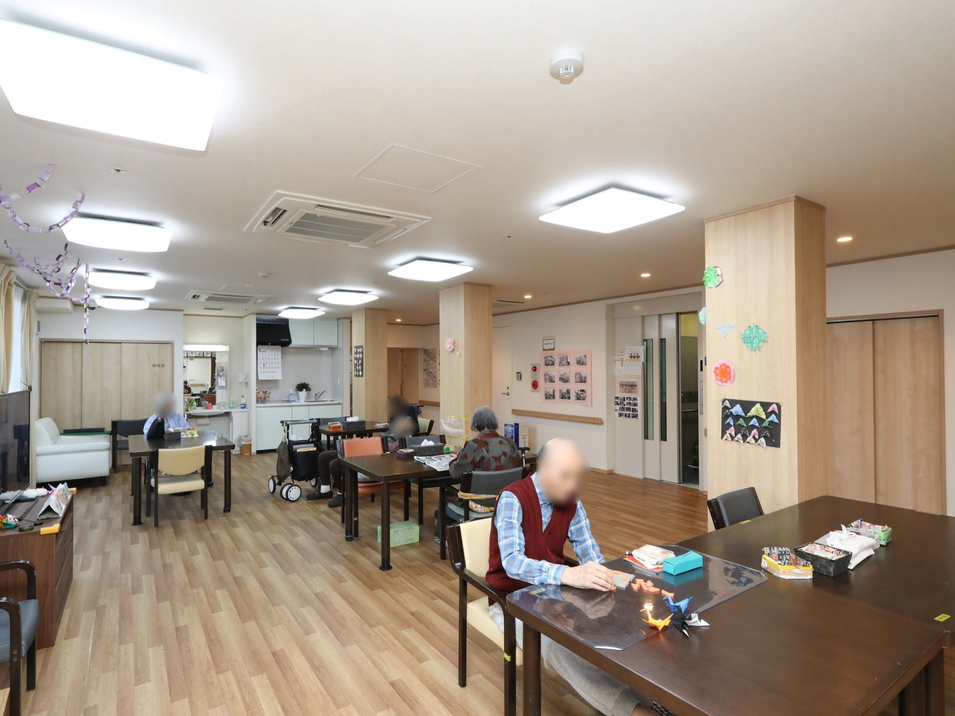 エレベーター乗降口の目の前に、広い共有スペースがある。多くの照明器具がついていてとても明るい。入居者様が４名着席している。
