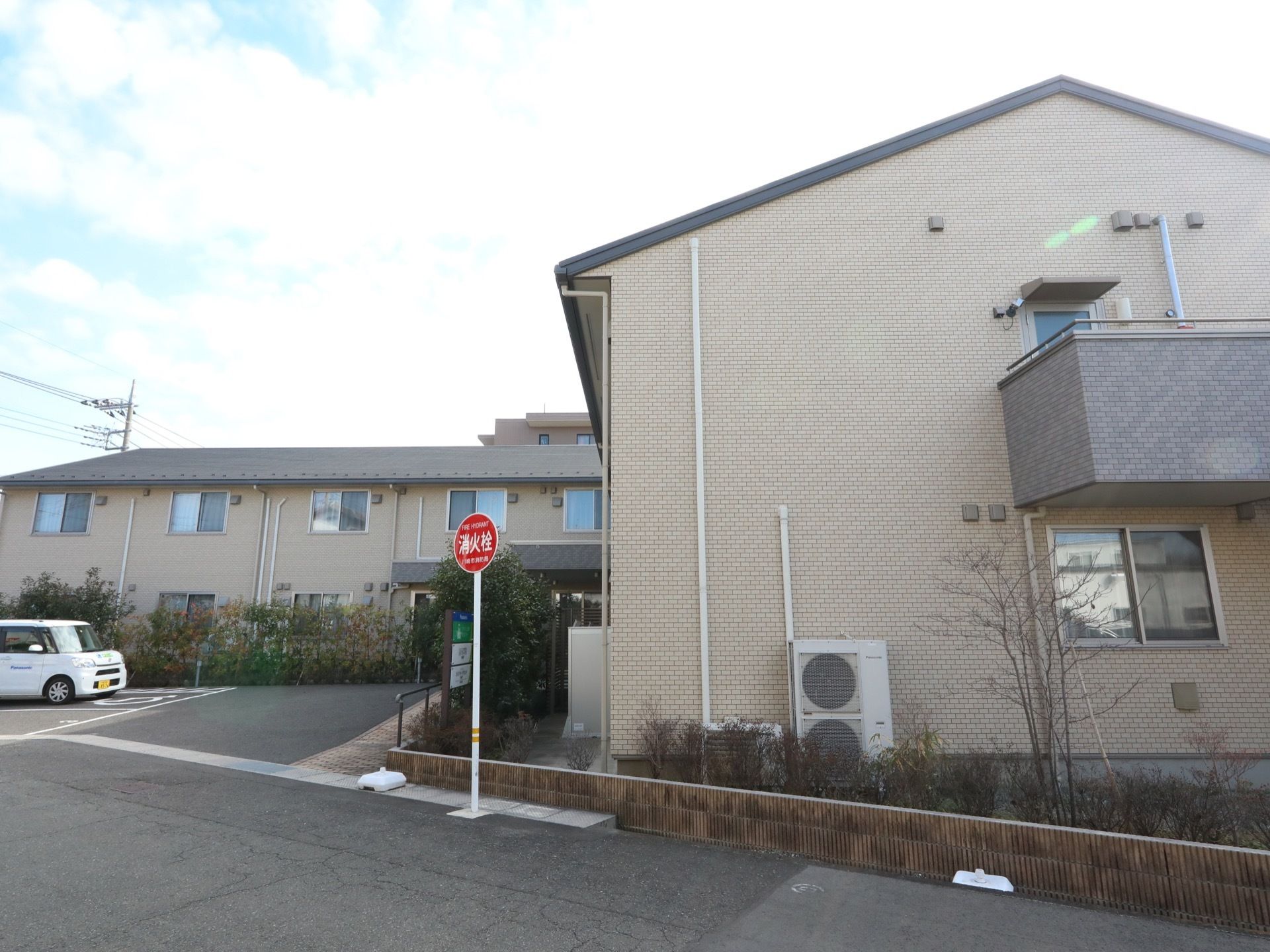 くの字型の構造をした２階建ての施設である。敷地内に白色の軽自動車が停まっている。敷地前の道路に消火栓の表示が立っている。