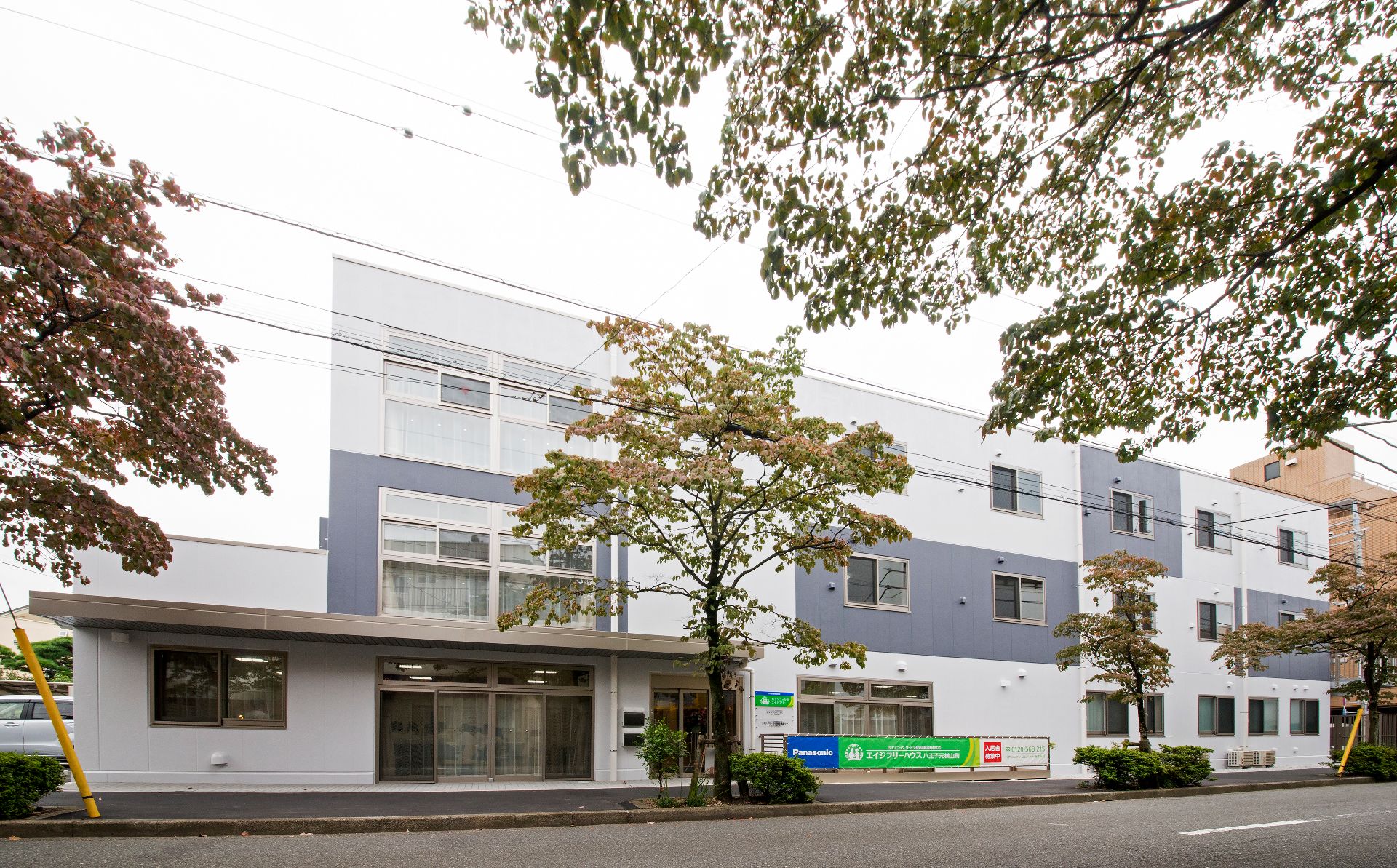 グレーと白の建物は3階建てで、玄関部分には屋根が付き、ウッドデッキも設置されている。