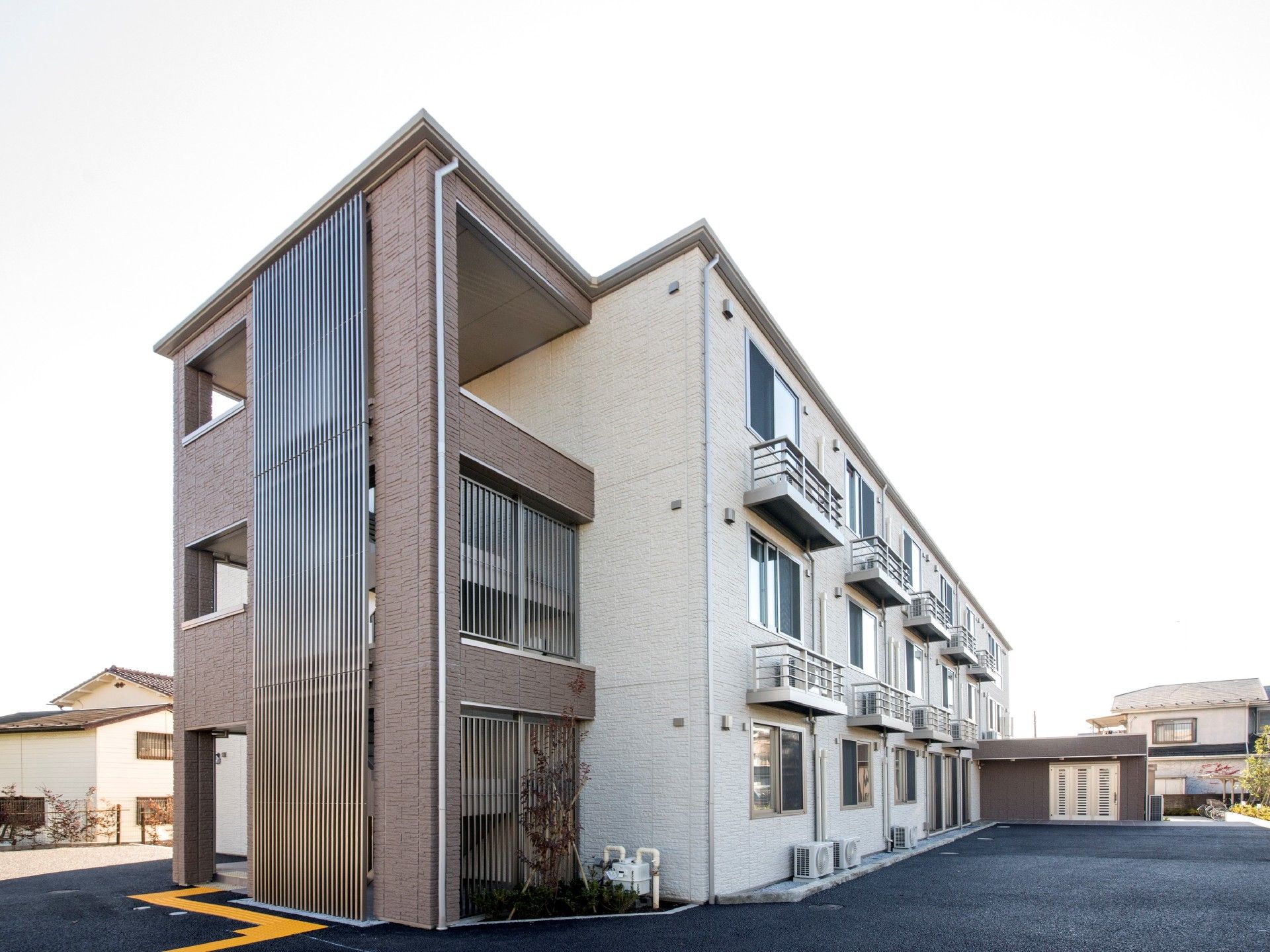 3階建ての建物はブラウン系で屋外階段が付き、周辺には住宅や自然がある。