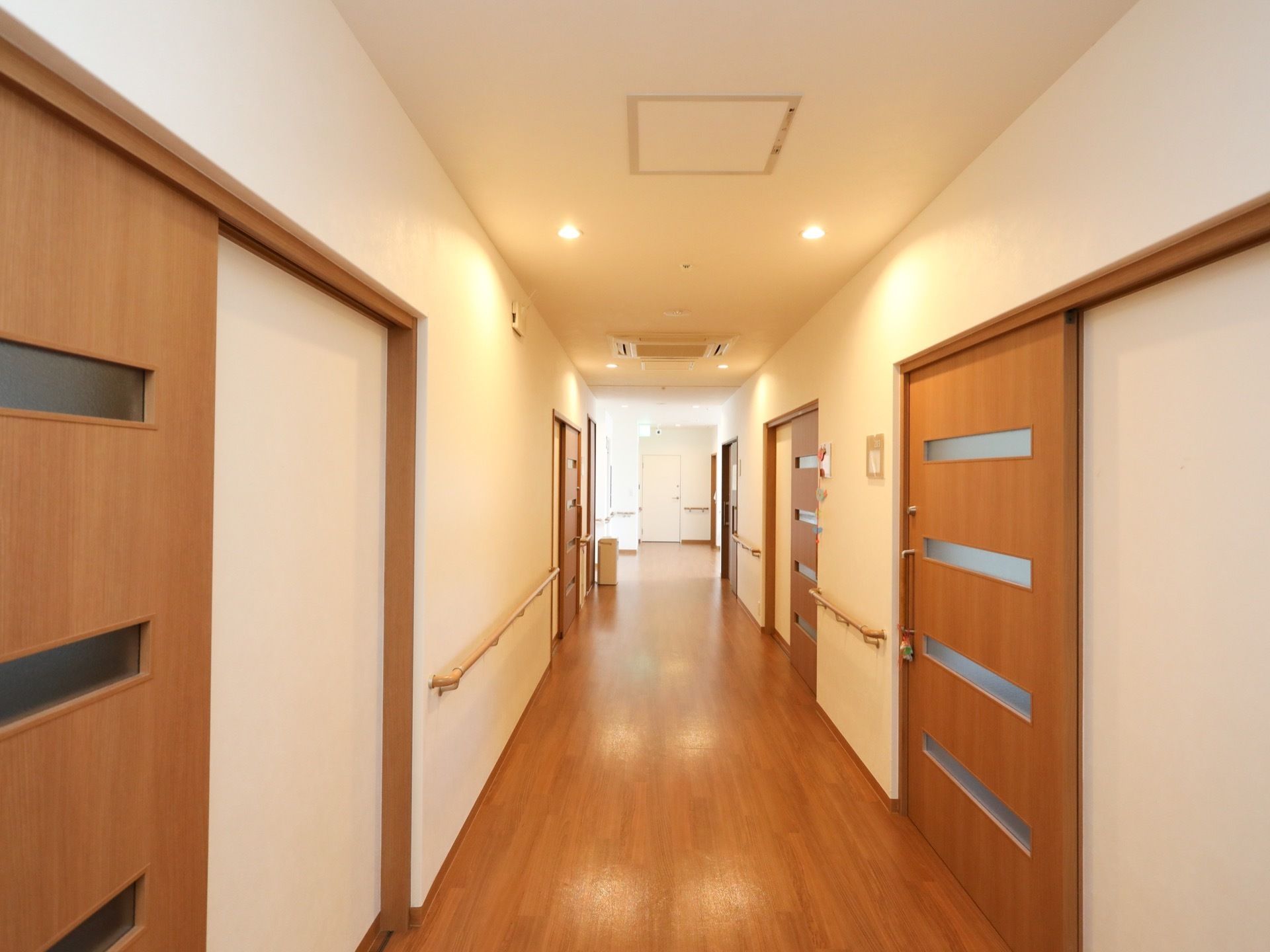 暖色系の照明が付けられた廊下で、壁には手すりが取り付けられている。居室のドアは木製で、すりガラスが等間隔にはめ込まれている。